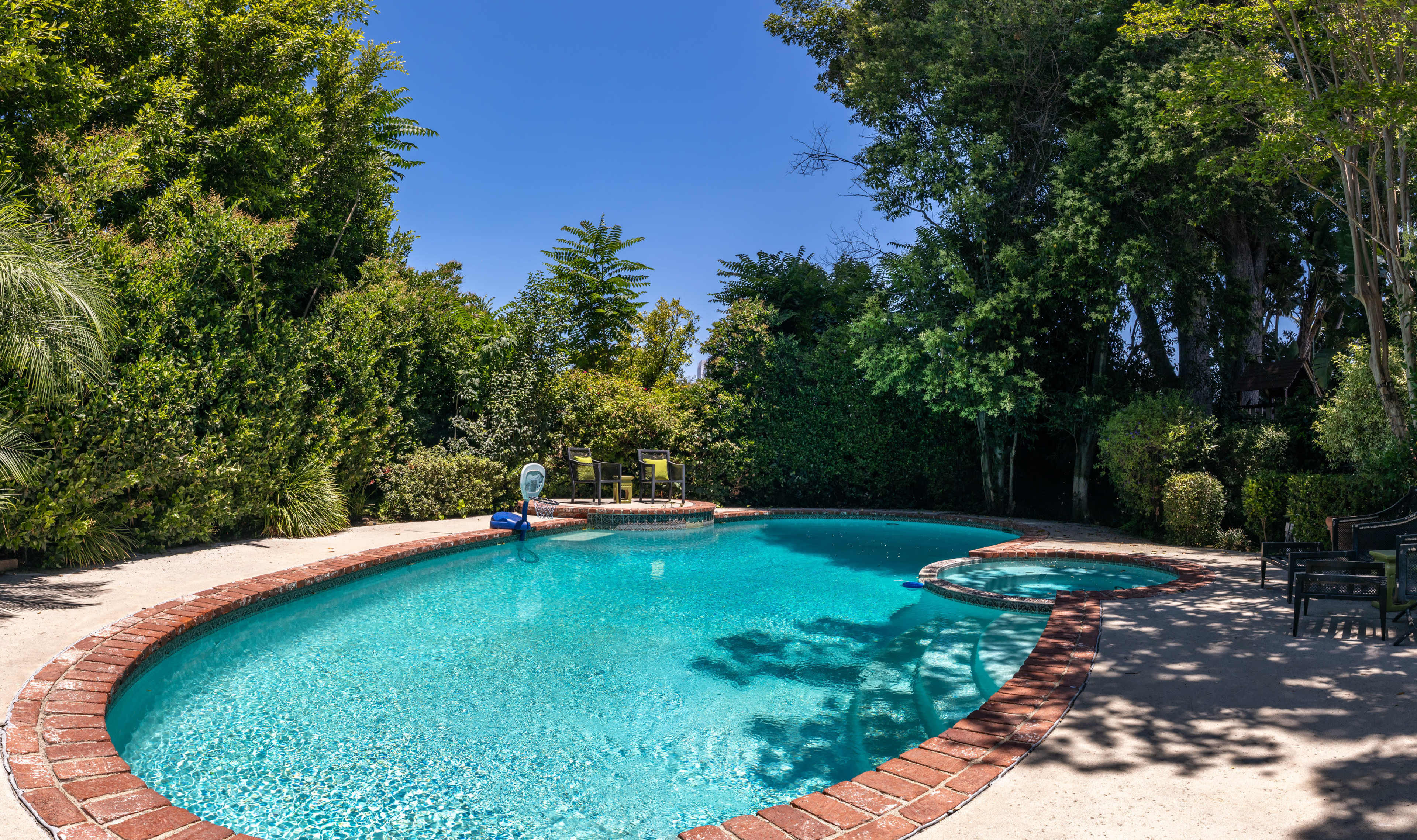 The image shows a clear swimming pool surrounded by lush greenery and a stone patio.