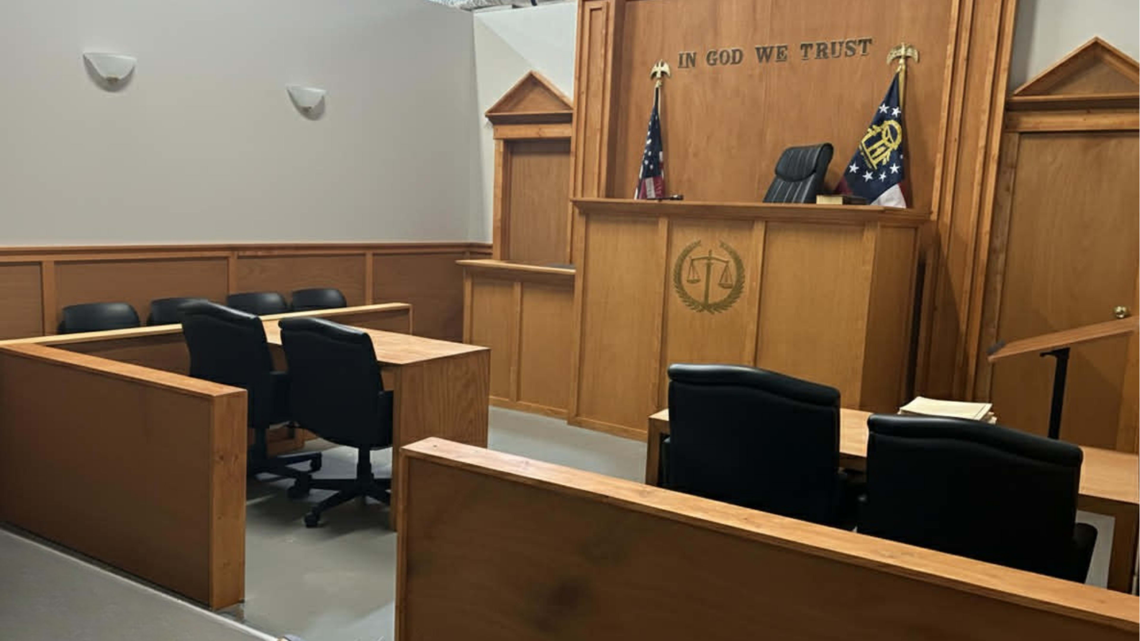 The image shows a courtroom interior with wooden benches, a judge's bench, and flags displayed in the background.
