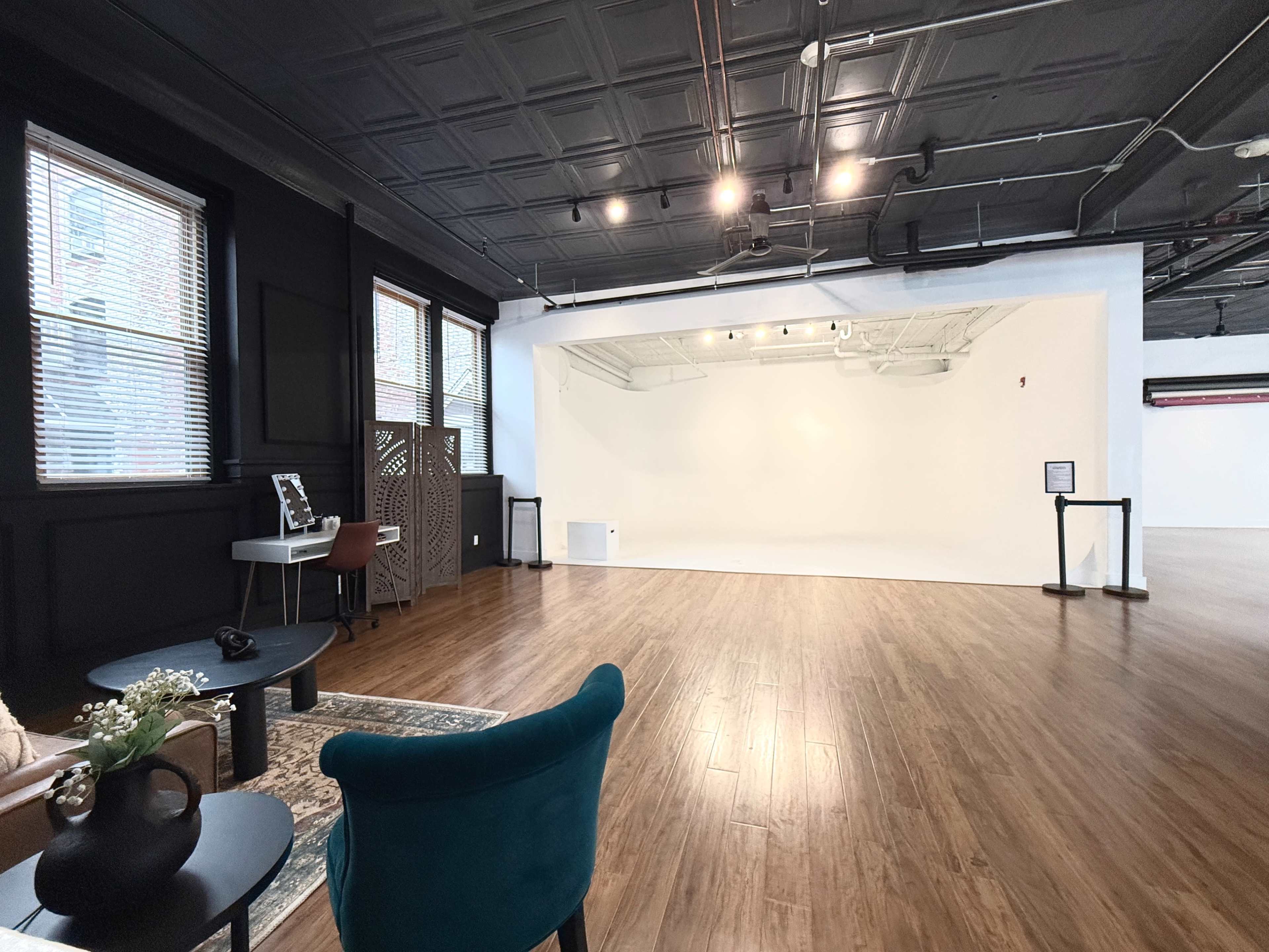 The image shows a spacious, minimally furnished studio with dark walls, polished wooden floors, and large windows allowing natural light.