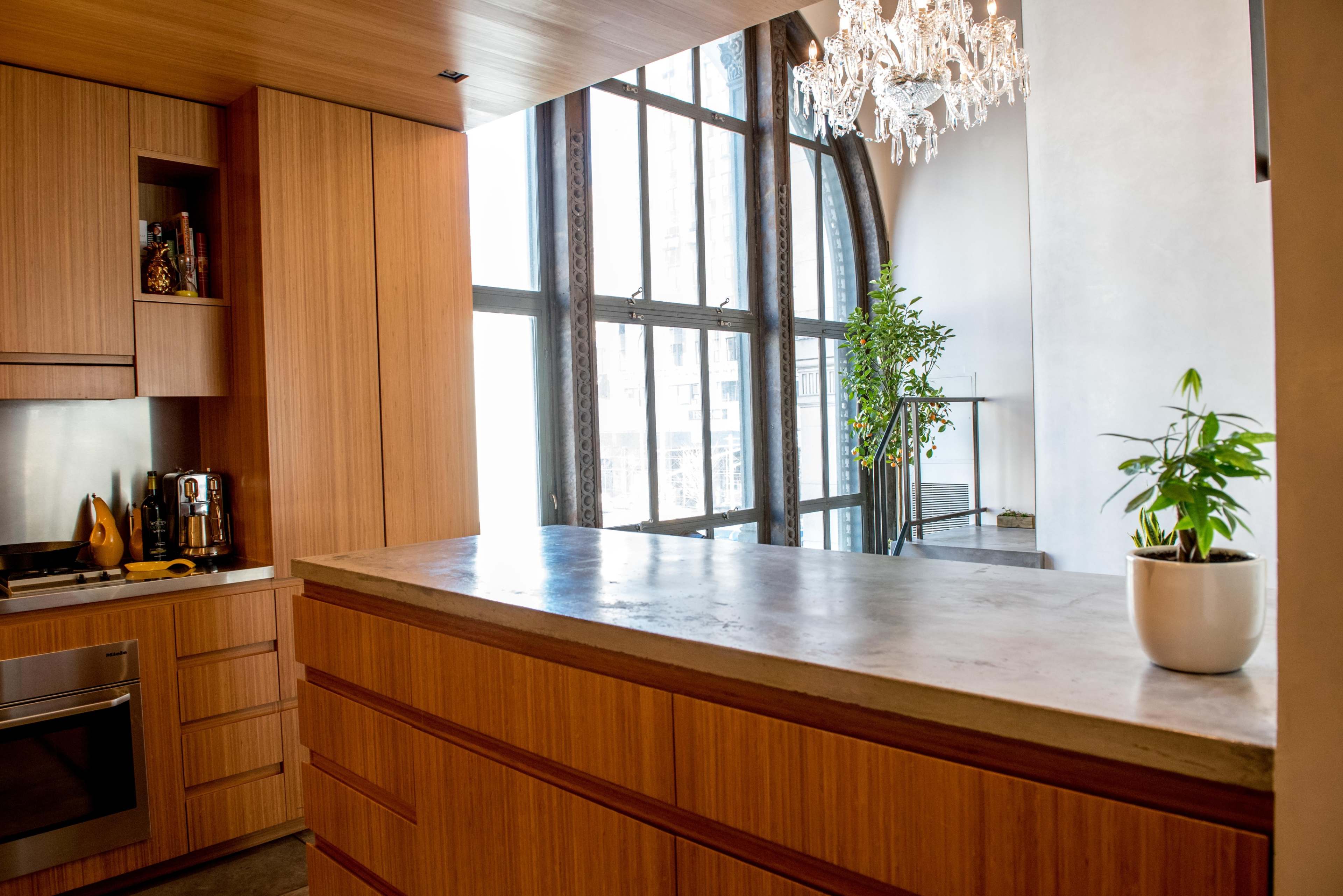 A modern kitchen features wooden cabinets, a concrete countertop, and large windows with a view of greenery outside.