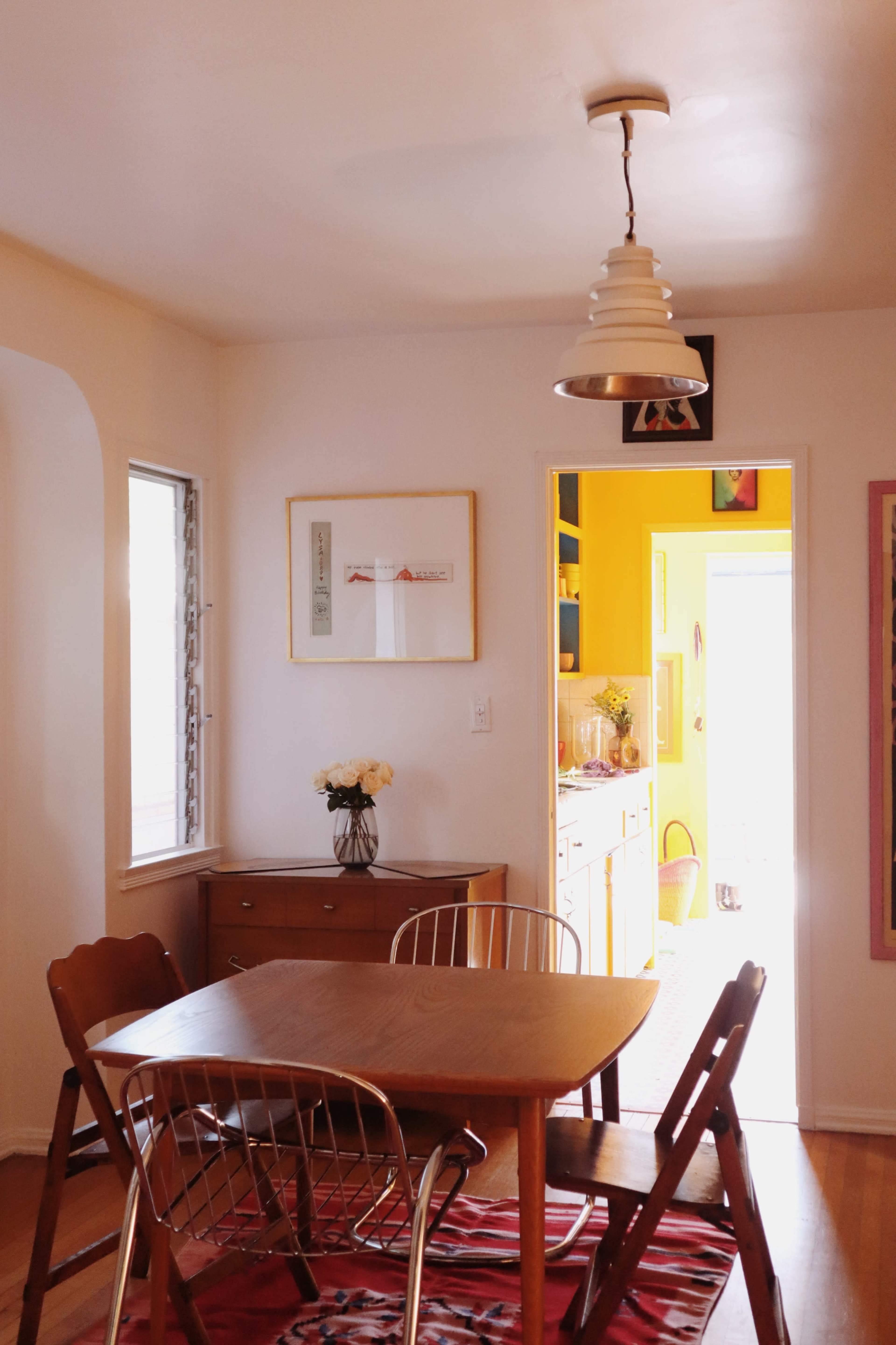 A dining area features a wooden table with metal chairs, a cabinet against the wall, and a doorway leading to a brightly colored kitchen.