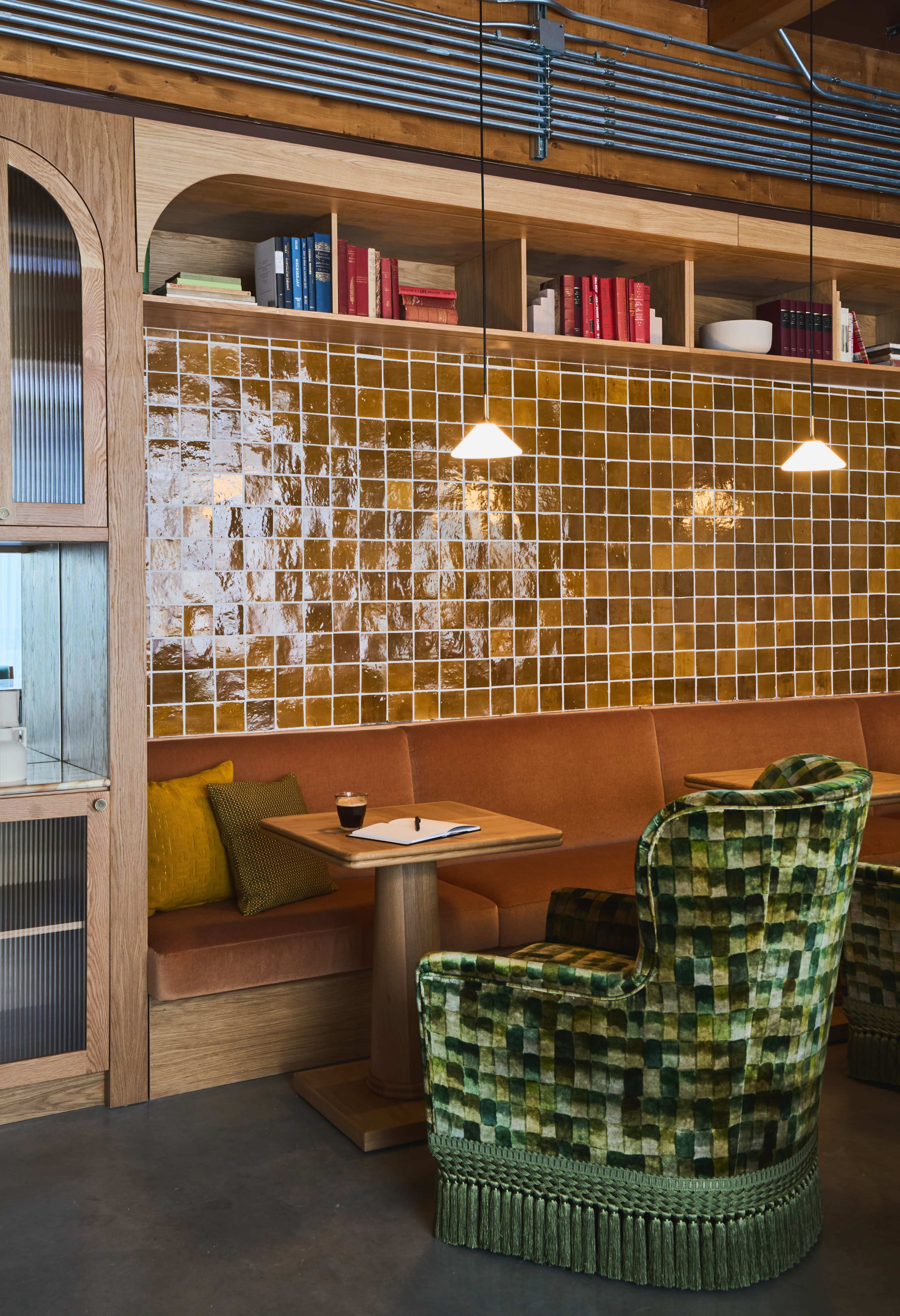 A modern café interior with an orange cushioned booth, green patterned chairs, and shelves filled with books and decorative items.