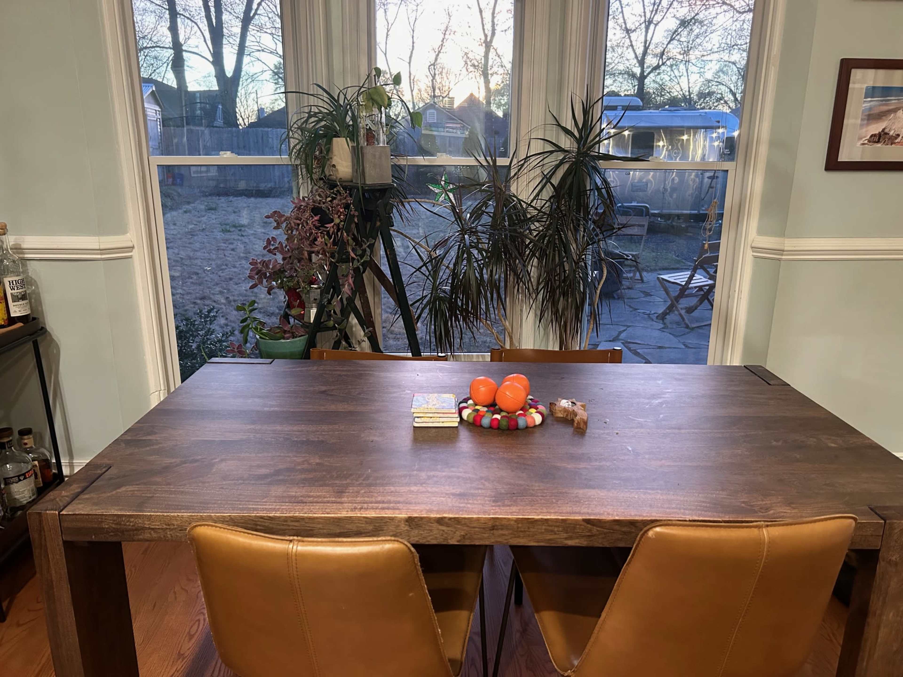 A wooden dining table with a colorful centerpiece of fruits is set in a room with large windows overlooking a garden.