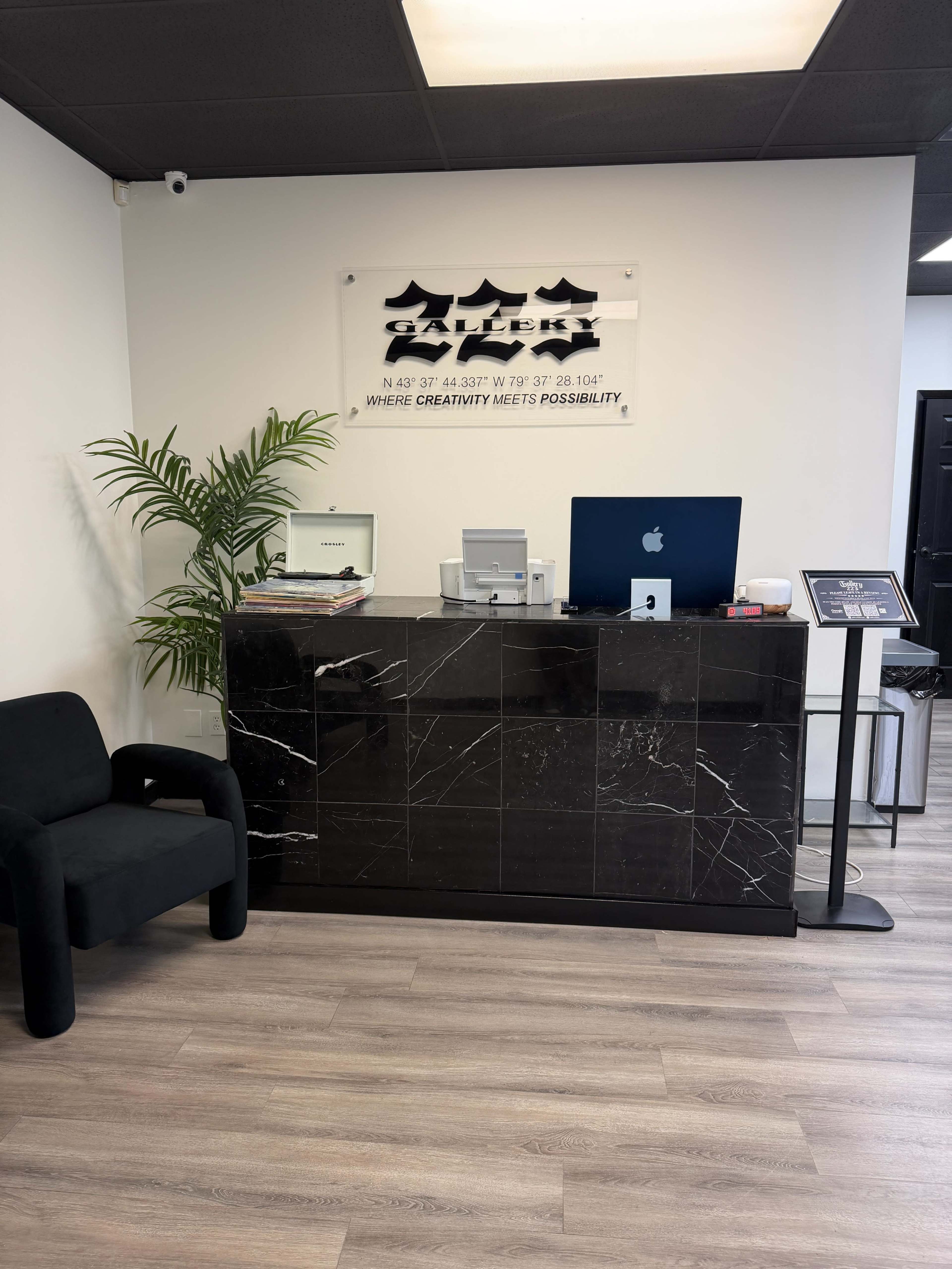 The image shows a modern reception desk made of dark marble with a computer and some magazines on it, beside a black chair and a wall featuring a sign with a creative slogan.