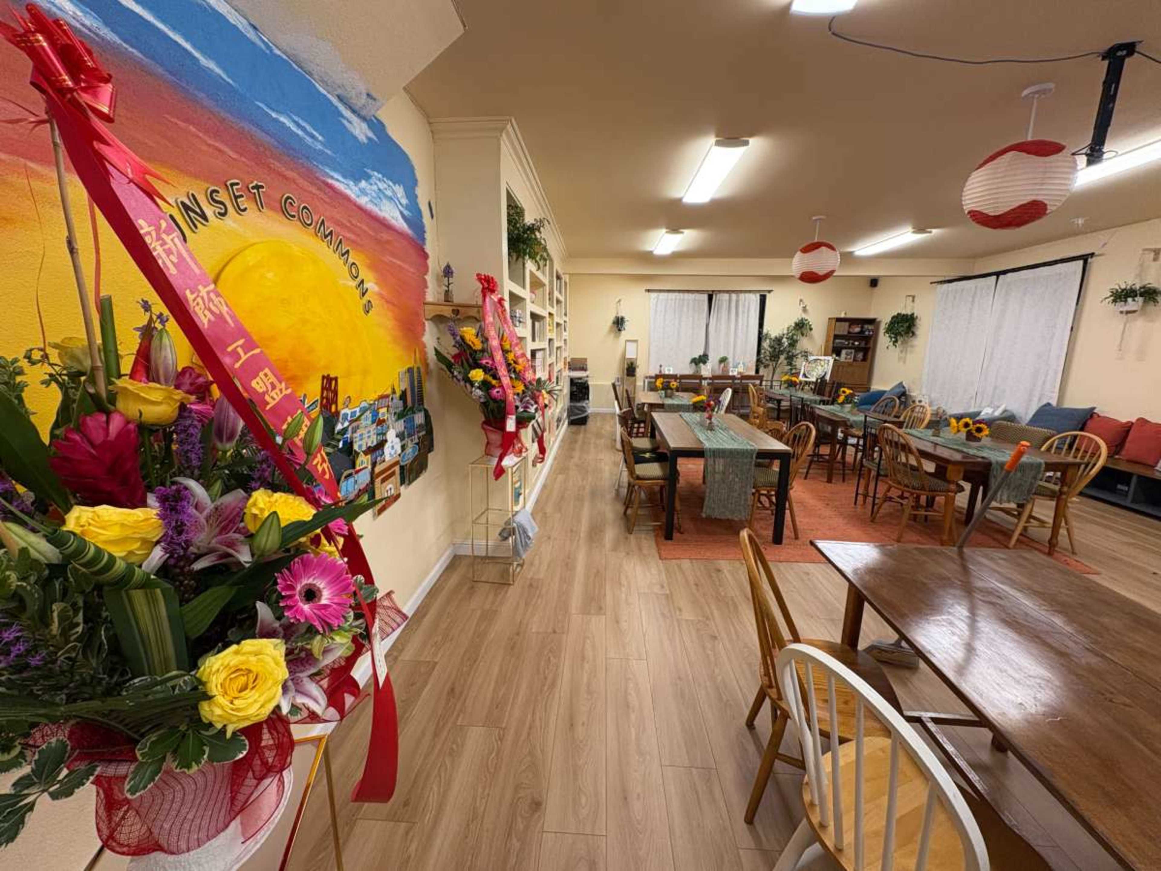 The image shows a bright and inviting communal space with large tables, colorful flower arrangements, and a mural on the wall.