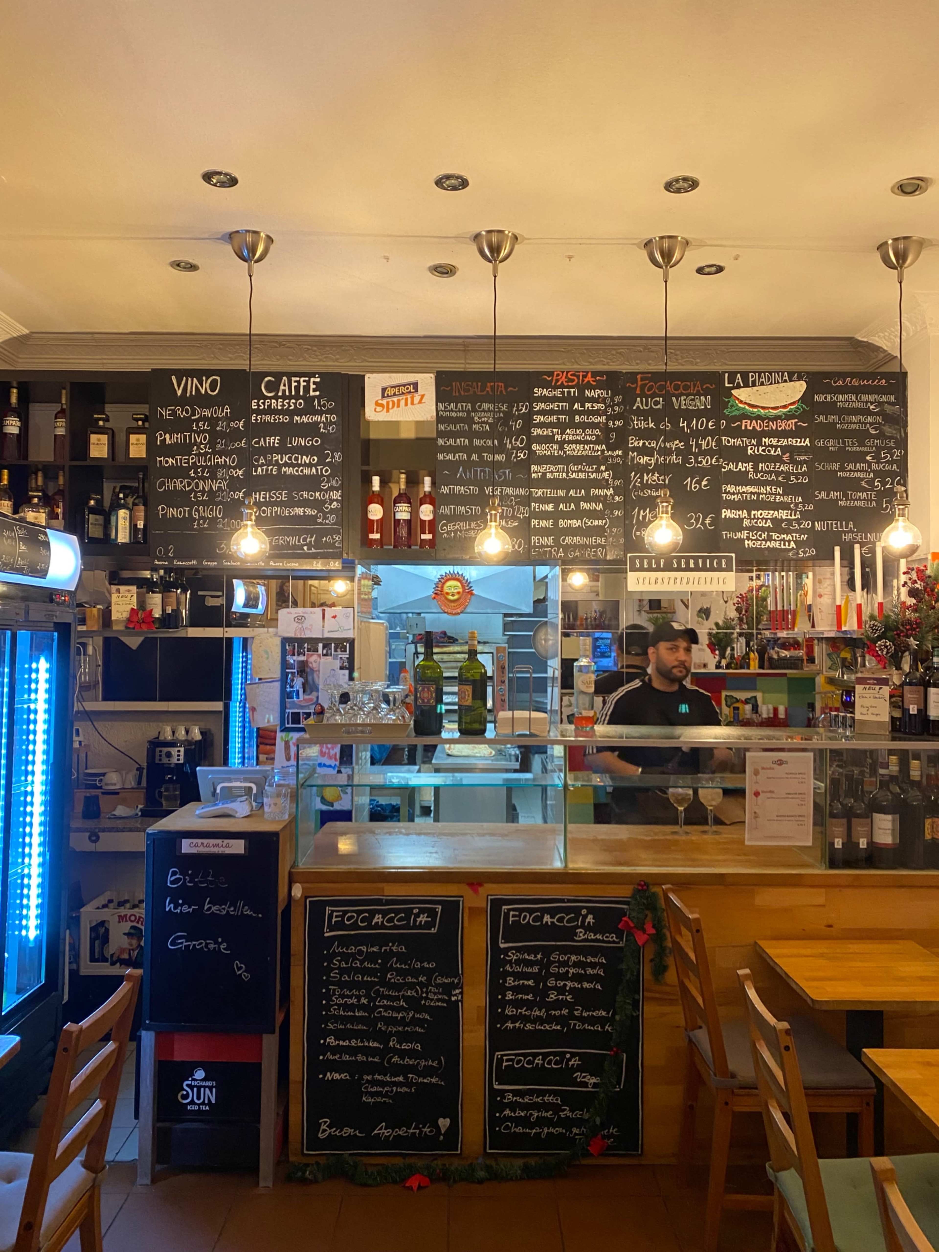 The image shows an interior of a small café with a bar area displaying a menu on the wall and various bottles on shelves.