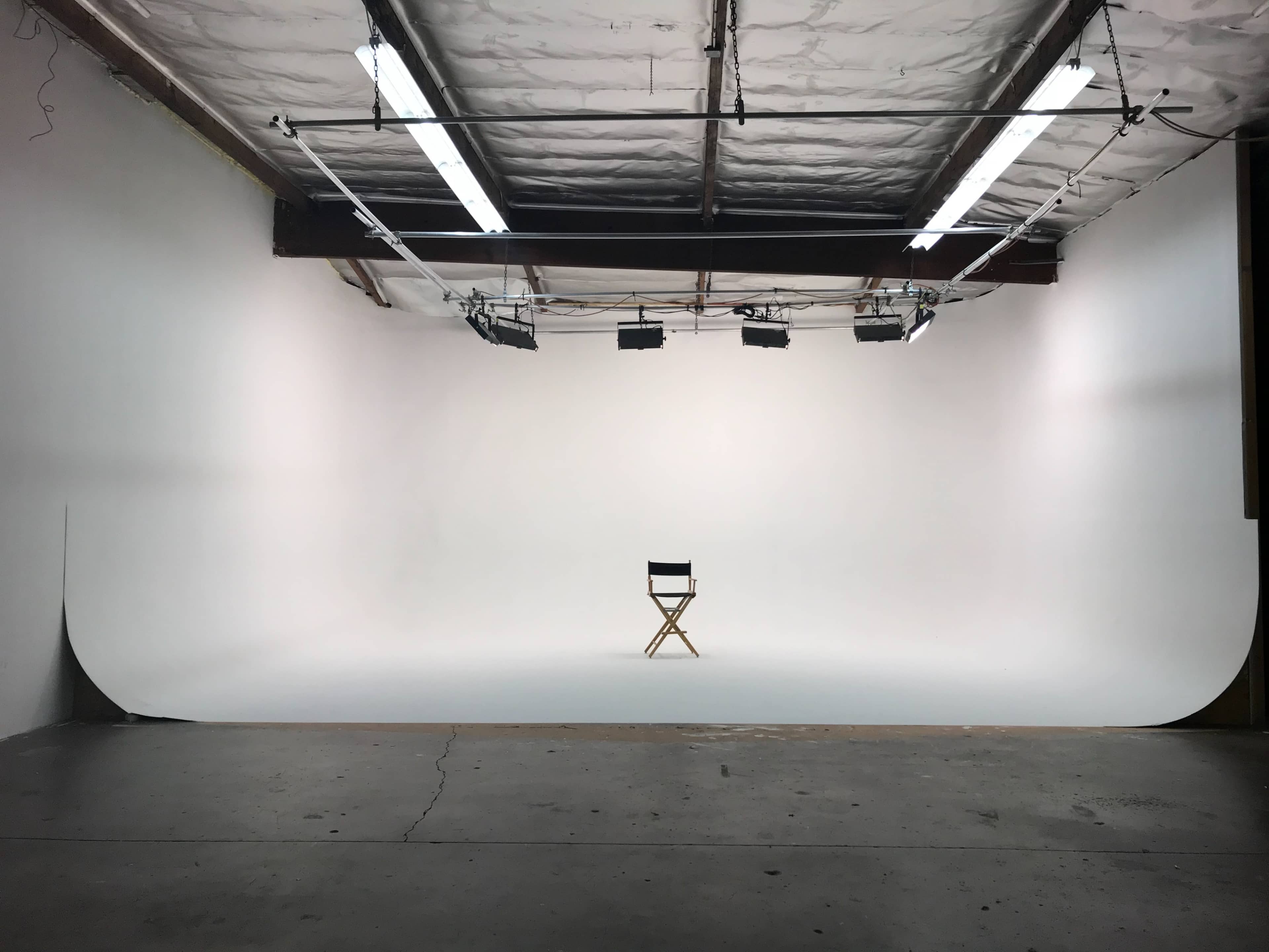 A wooden director's chair stands alone in the center of a spacious, white photography studio.
