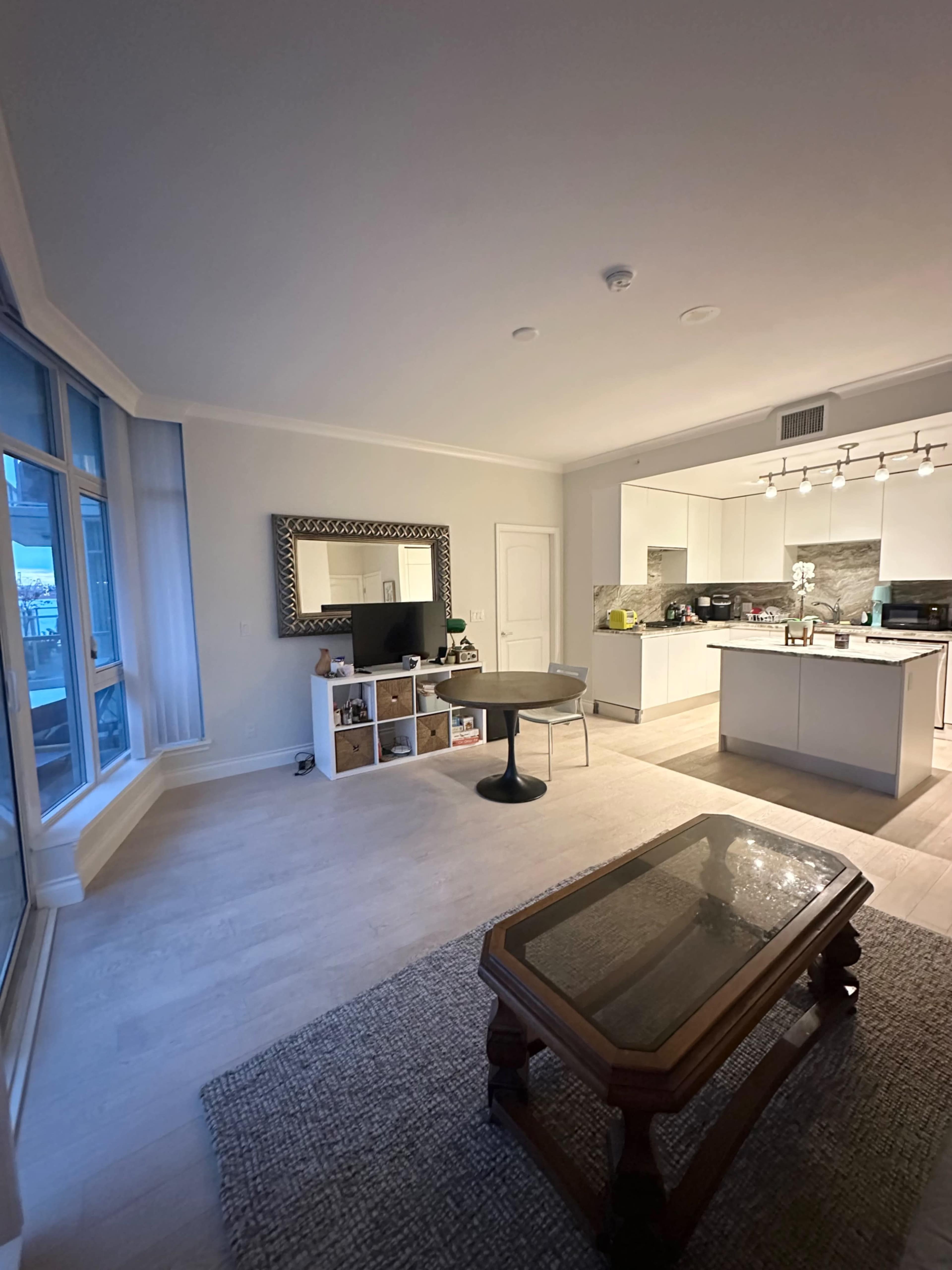A modern, minimally furnished living space features a circular dining table, a glass display coffee table, and a kitchen area with sleek cabinets and a large mirror on the wall.