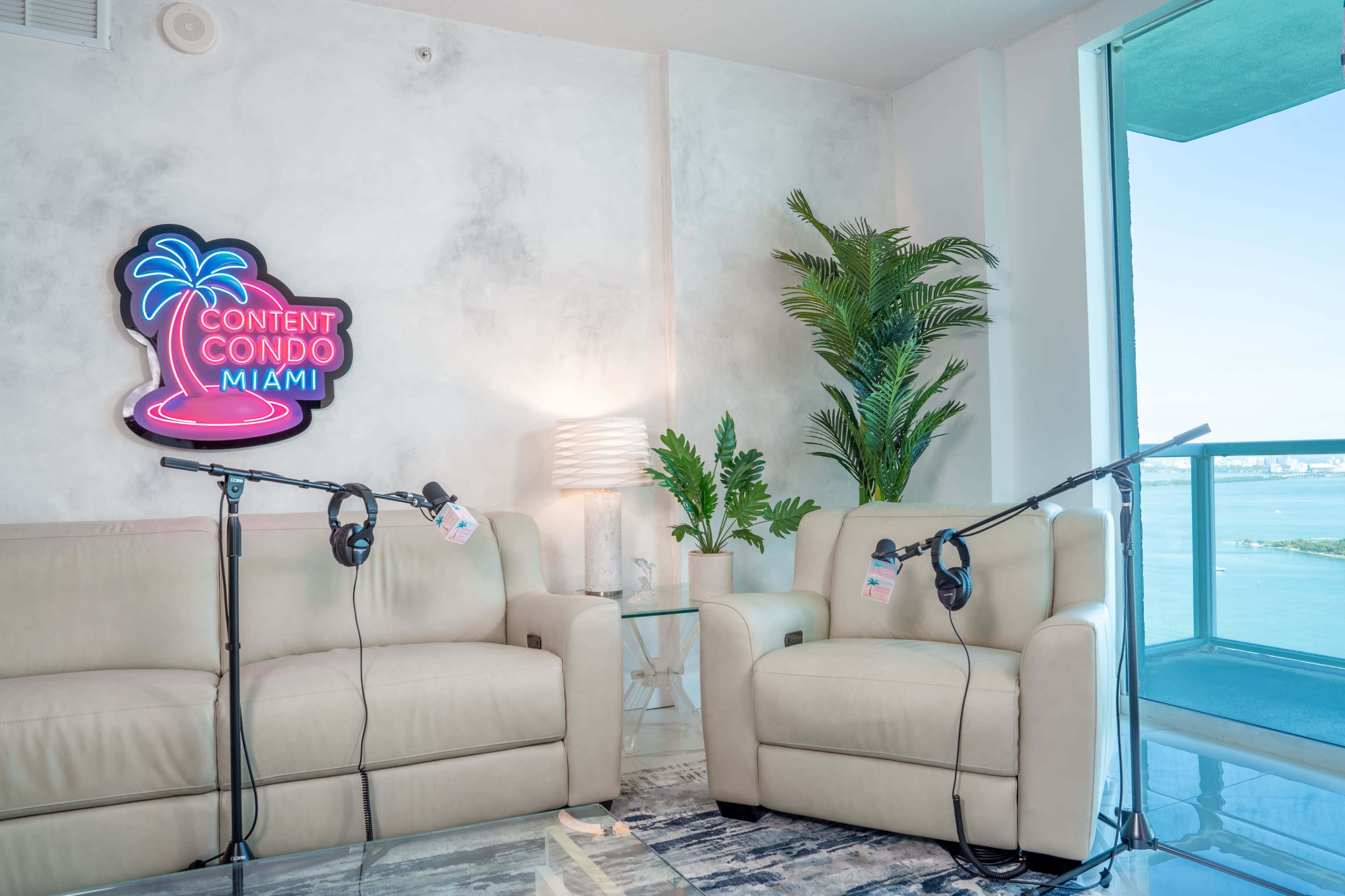 The image shows a modern room with two beige sofas, a neon sign that reads "Content Condo Miami," and a view of water through a large window.