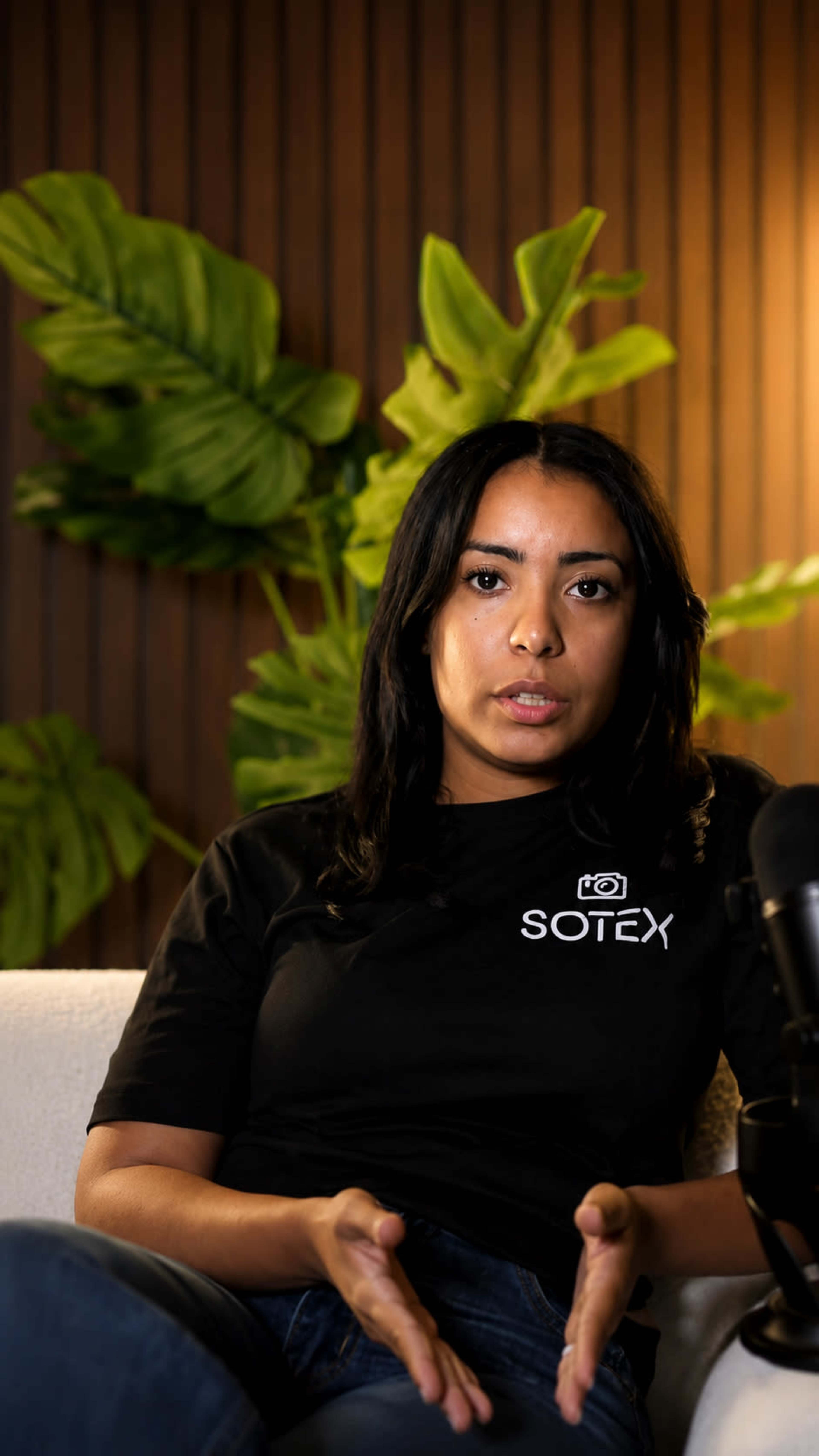 A woman in a black SOTEX t-shirt is sitting on a couch, gesturing with her hands while speaking in front of a backdrop of large green leaves.