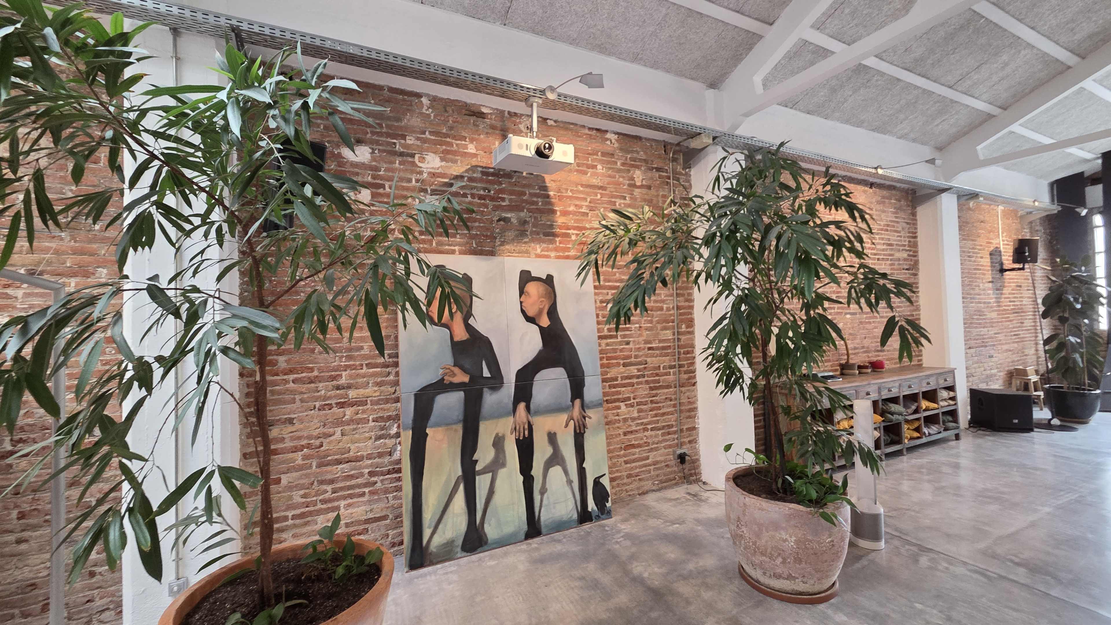 The image shows a spacious room with exposed brick walls, two large potted plants, and a painting of two figures centered on the wall.