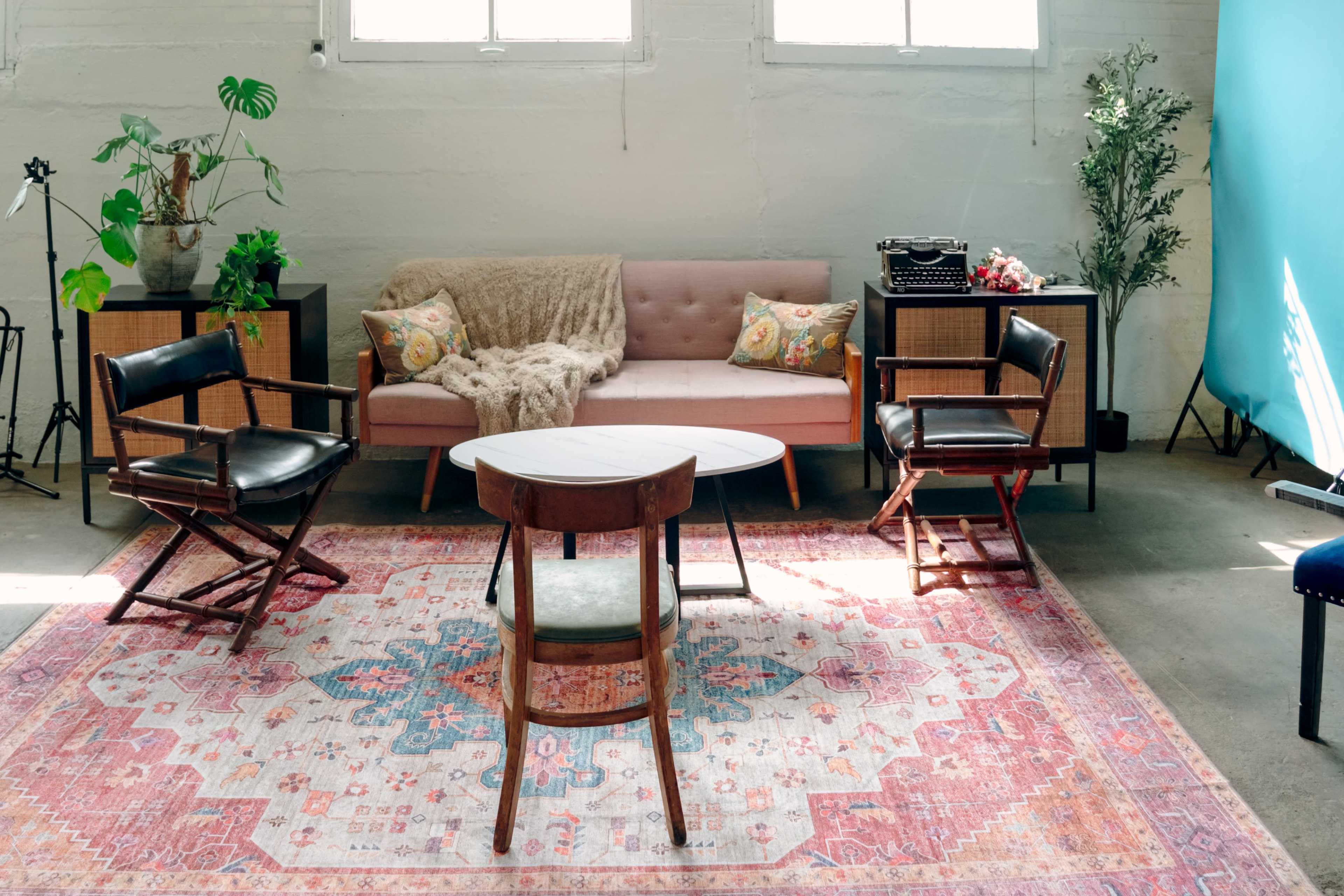 The image depicts a cozy interior space featuring a pink sofa, two wooden chairs, a round table, and decorative plants, all set on a patterned rug.