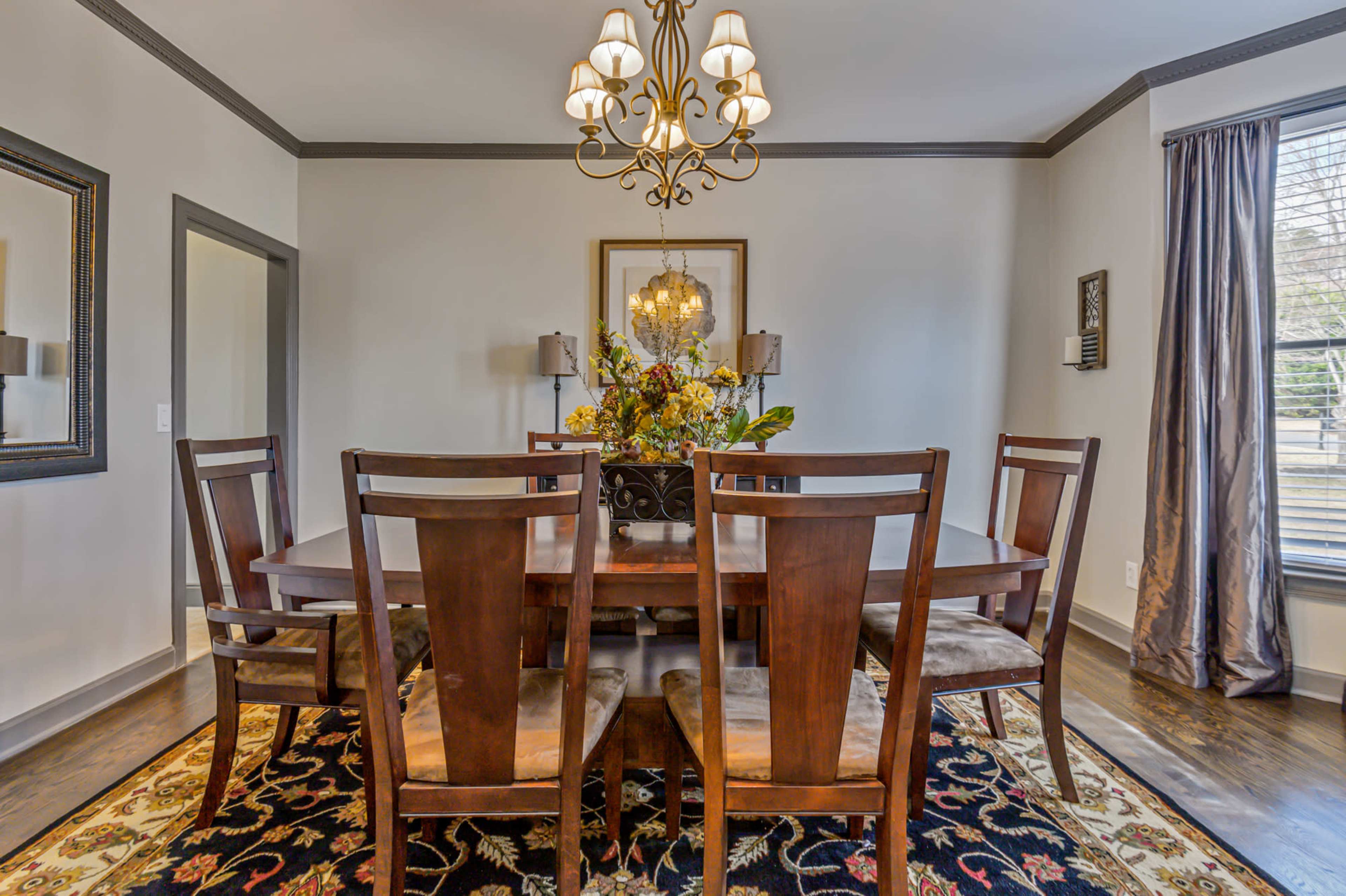 The image shows a dining room with a wooden table surrounded by six chairs, a chandelier above, and a floral centerpiece.