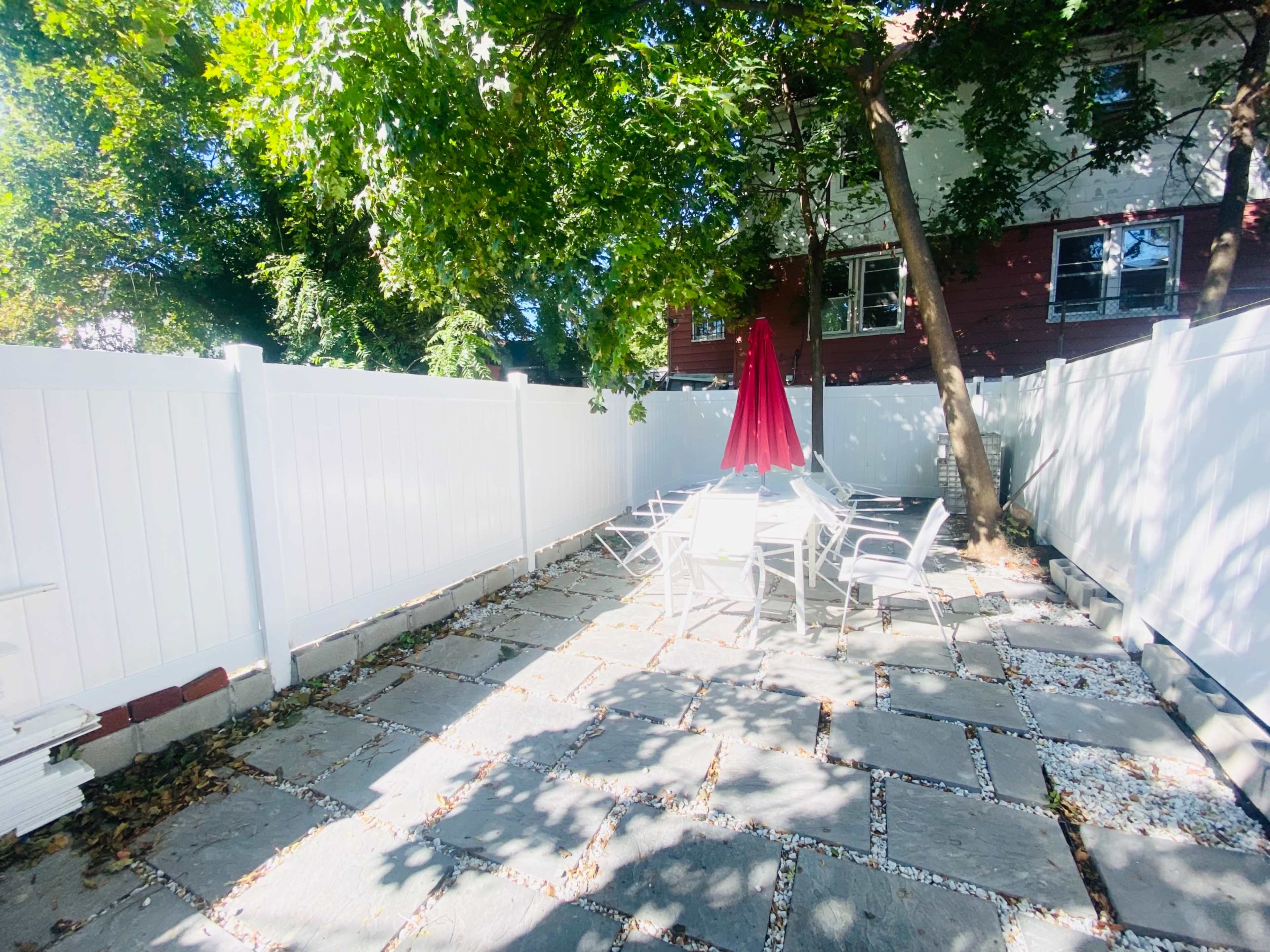 A fenced patio area features a table and chairs shaded by a red umbrella, surrounded by trees and a white fence.