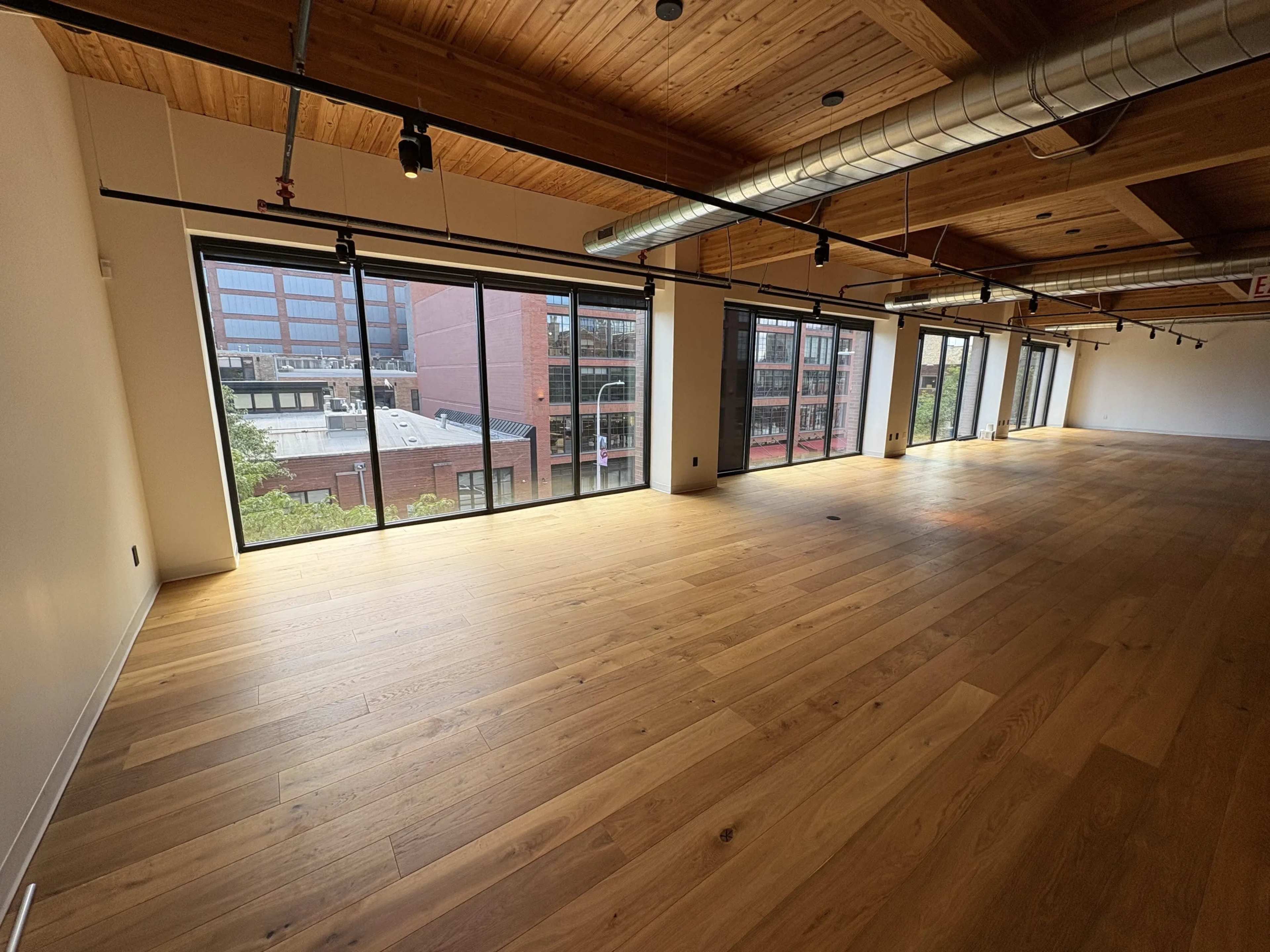 The image shows an empty room with wooden flooring and large windows, providing a view of adjacent buildings.