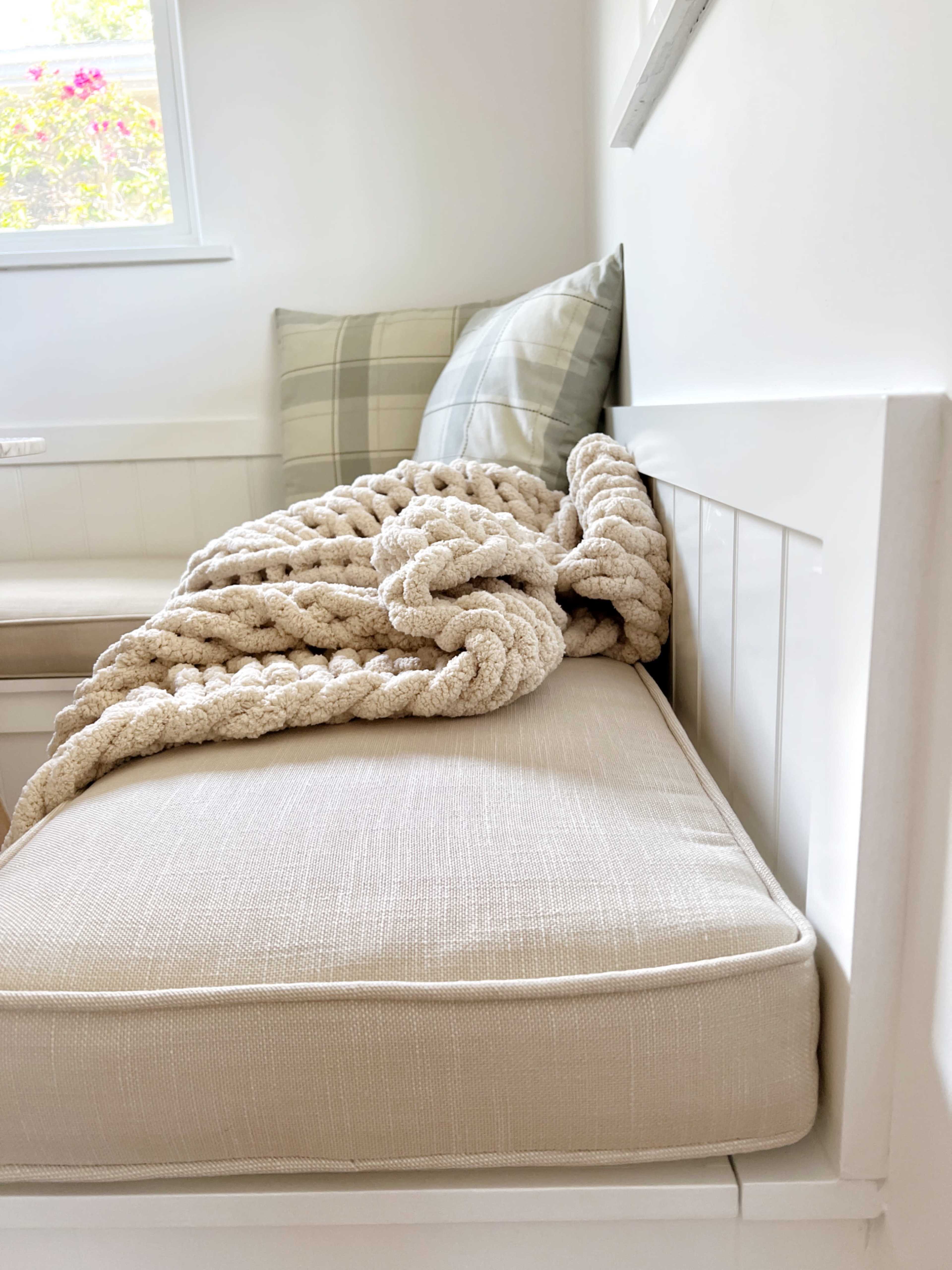 A beige couch with a thick, textured blanket draped over it is positioned against a white wall in a brightly lit room.