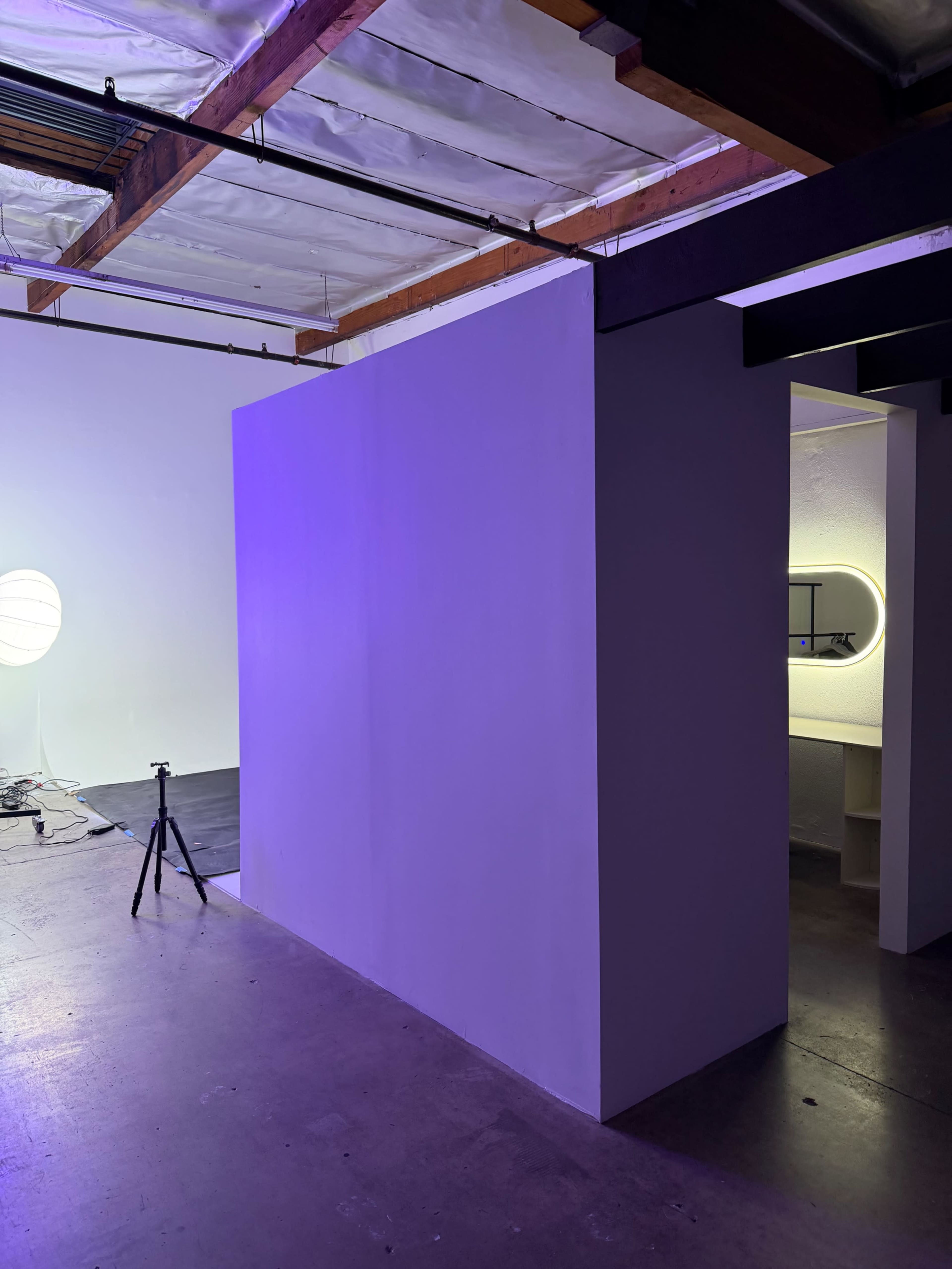 The image shows a partially painted room with a white wall and a small, illuminated section featuring an arched mirror and a tripod set up for photography.