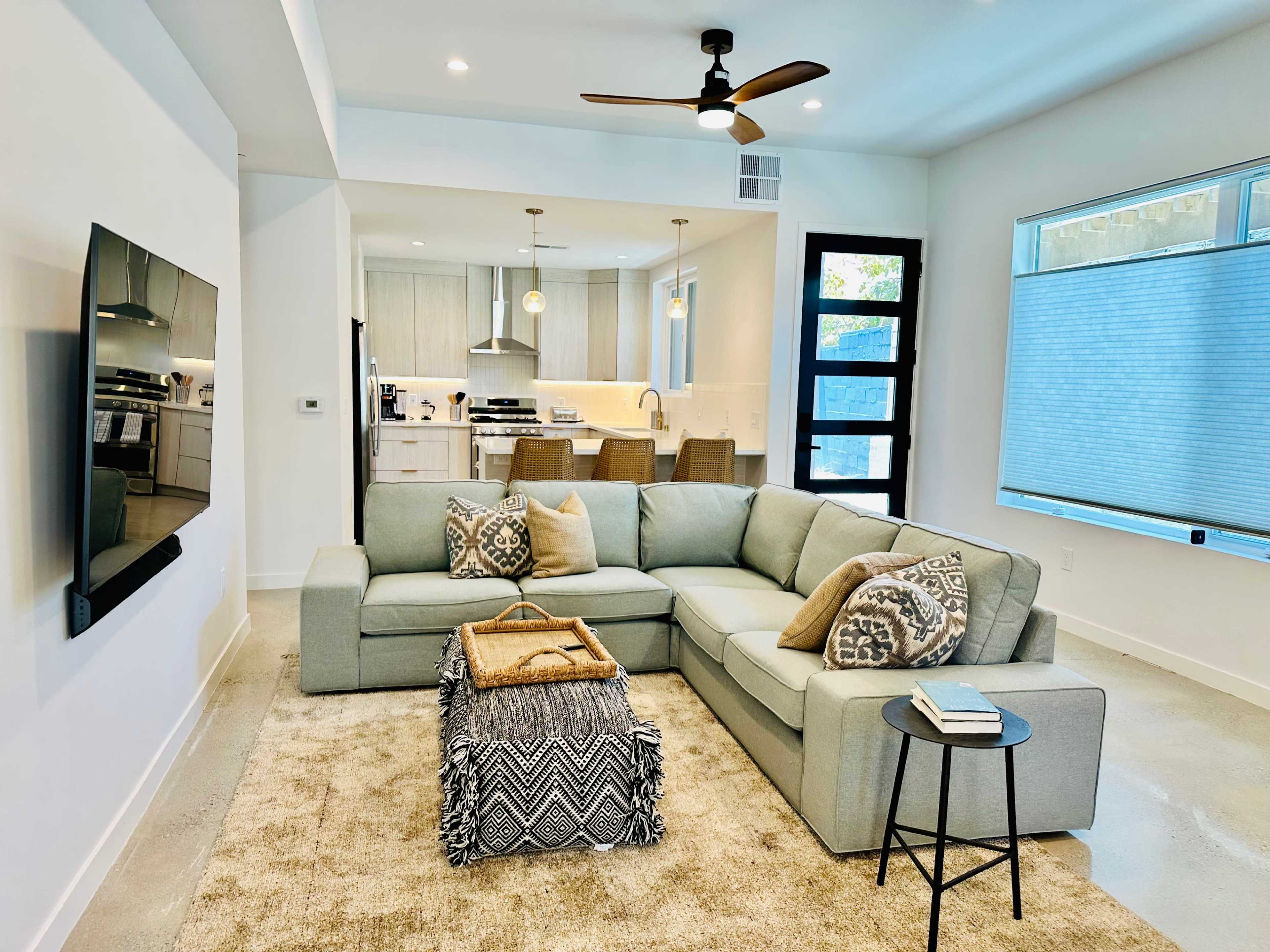 The image shows a modern living room with a gray sectional sofa, a patterned throw blanket, and a small side table, leading into a kitchen area with light cabinetry and bar seating.