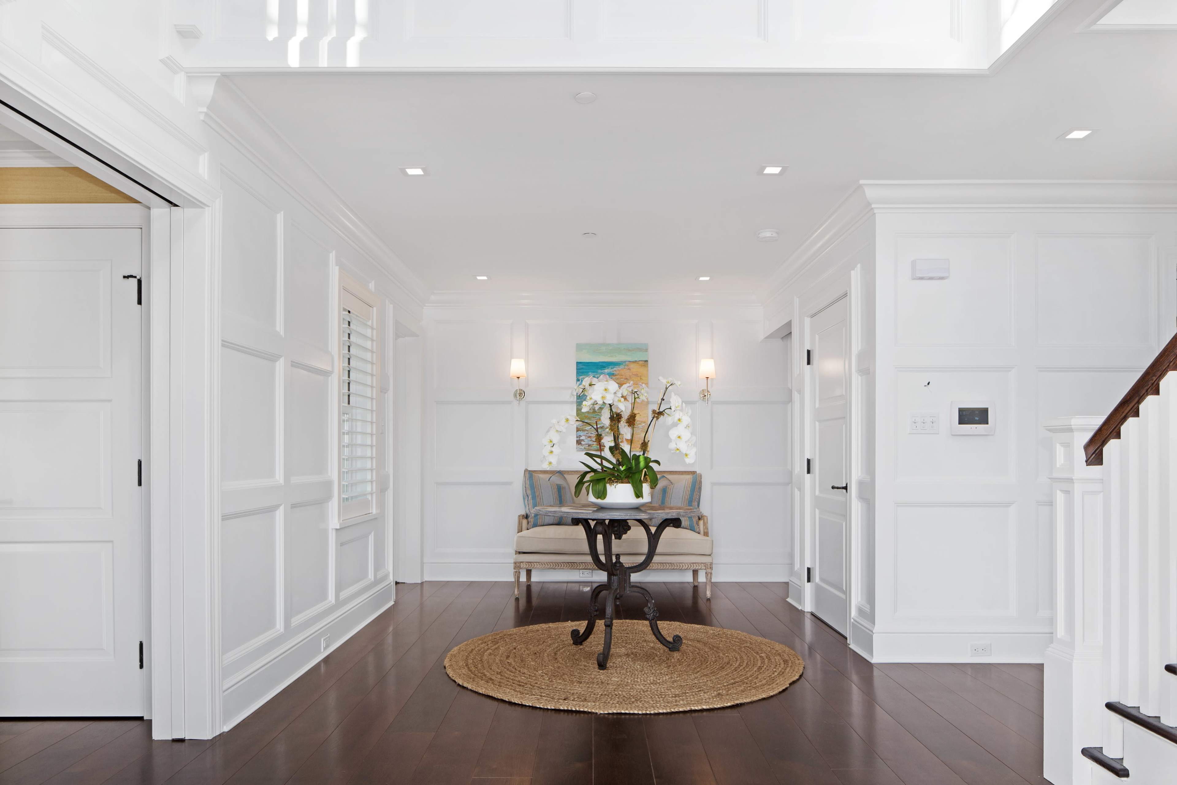 A bright foyer with white paneled walls, a round woven rug, and a small table topped with a bouquet of orchids.