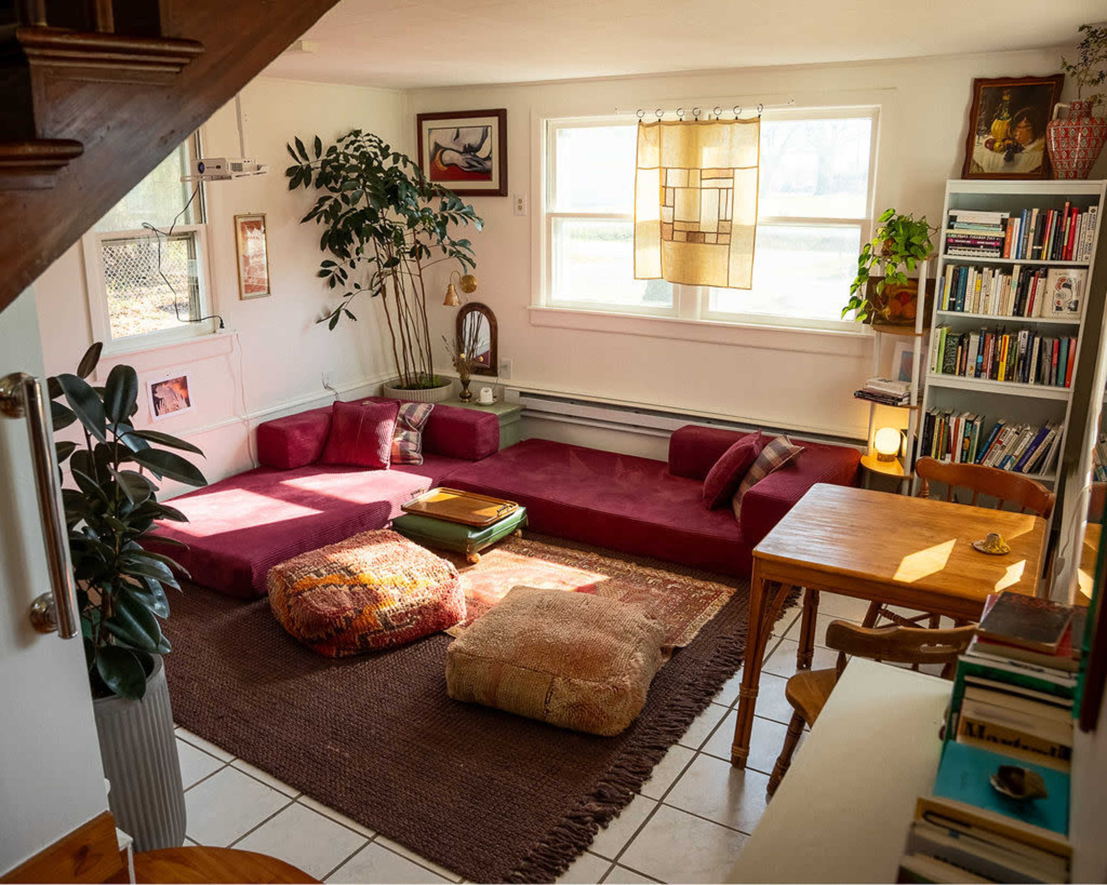 The image shows a cozy living space with a maroon seating area, a wooden table, bookshelves, and plants, illuminated by natural light from a window.