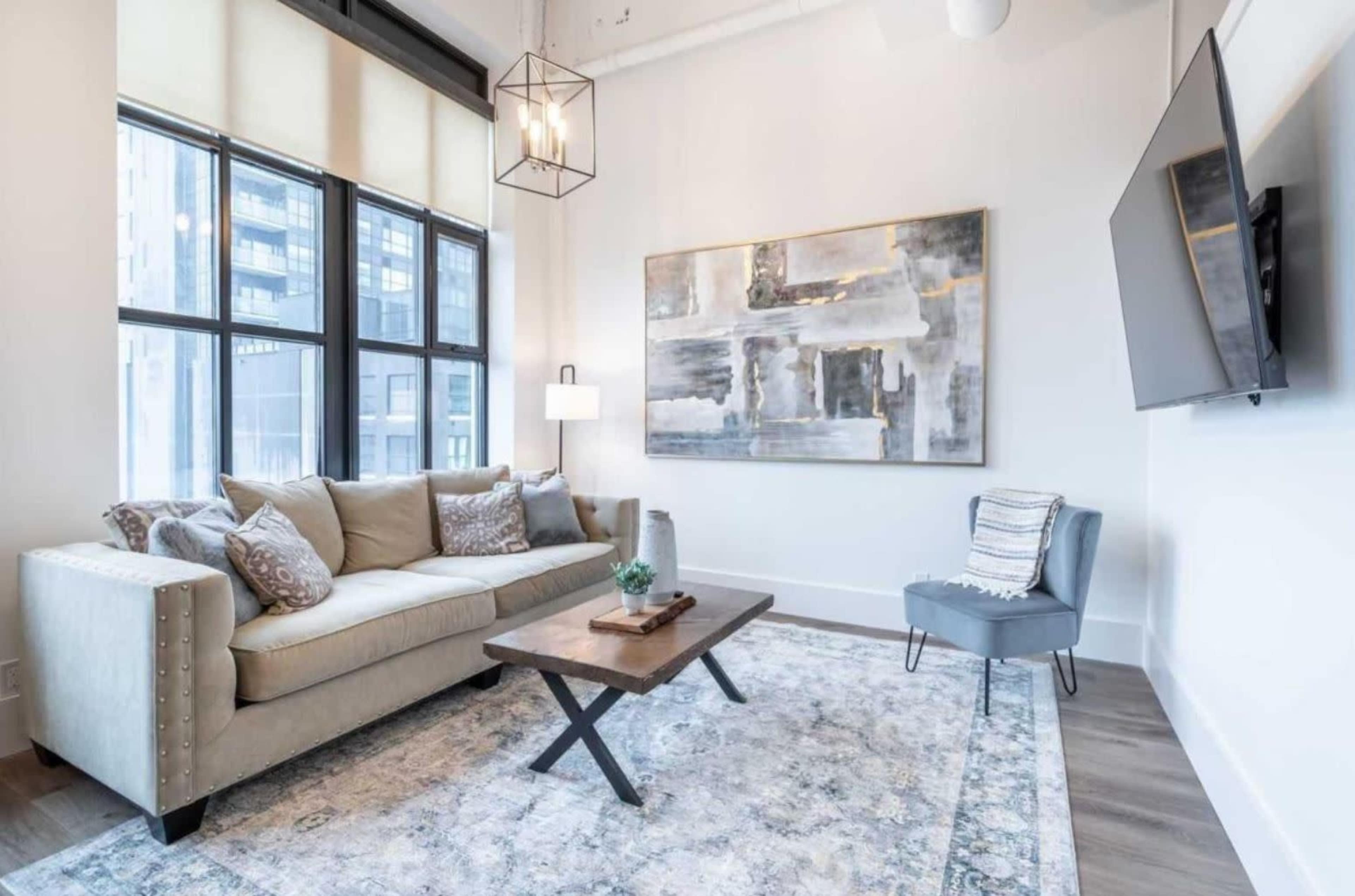 A modern living room with a beige sofa, a coffee table, and a small armchair, set against large windows and adorned with a piece of abstract art on the wall.