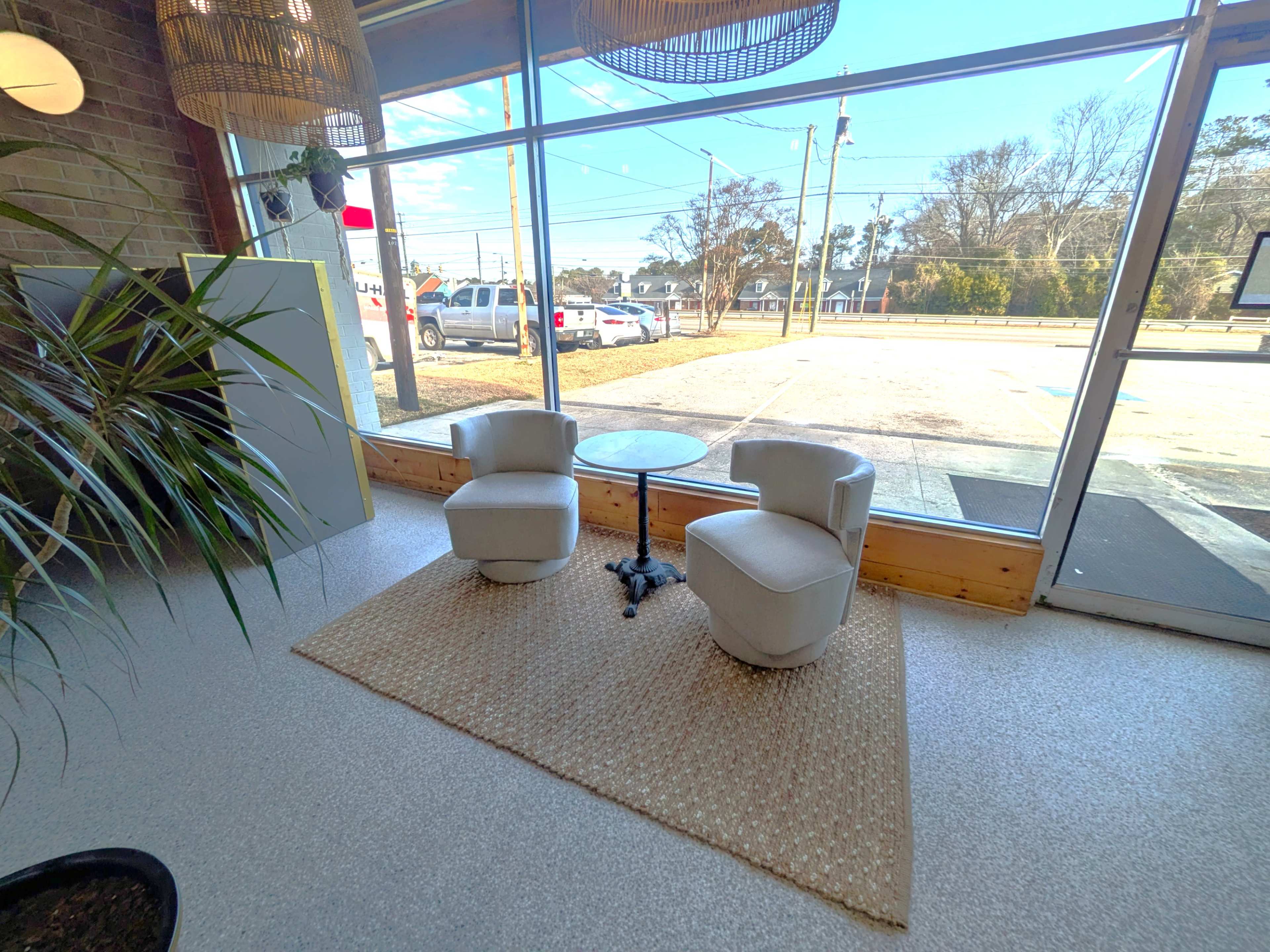 The image shows a cozy seating area with two white armchairs and a small round table on a beige rug, situated in front of a large window that offers a view of the outside.
