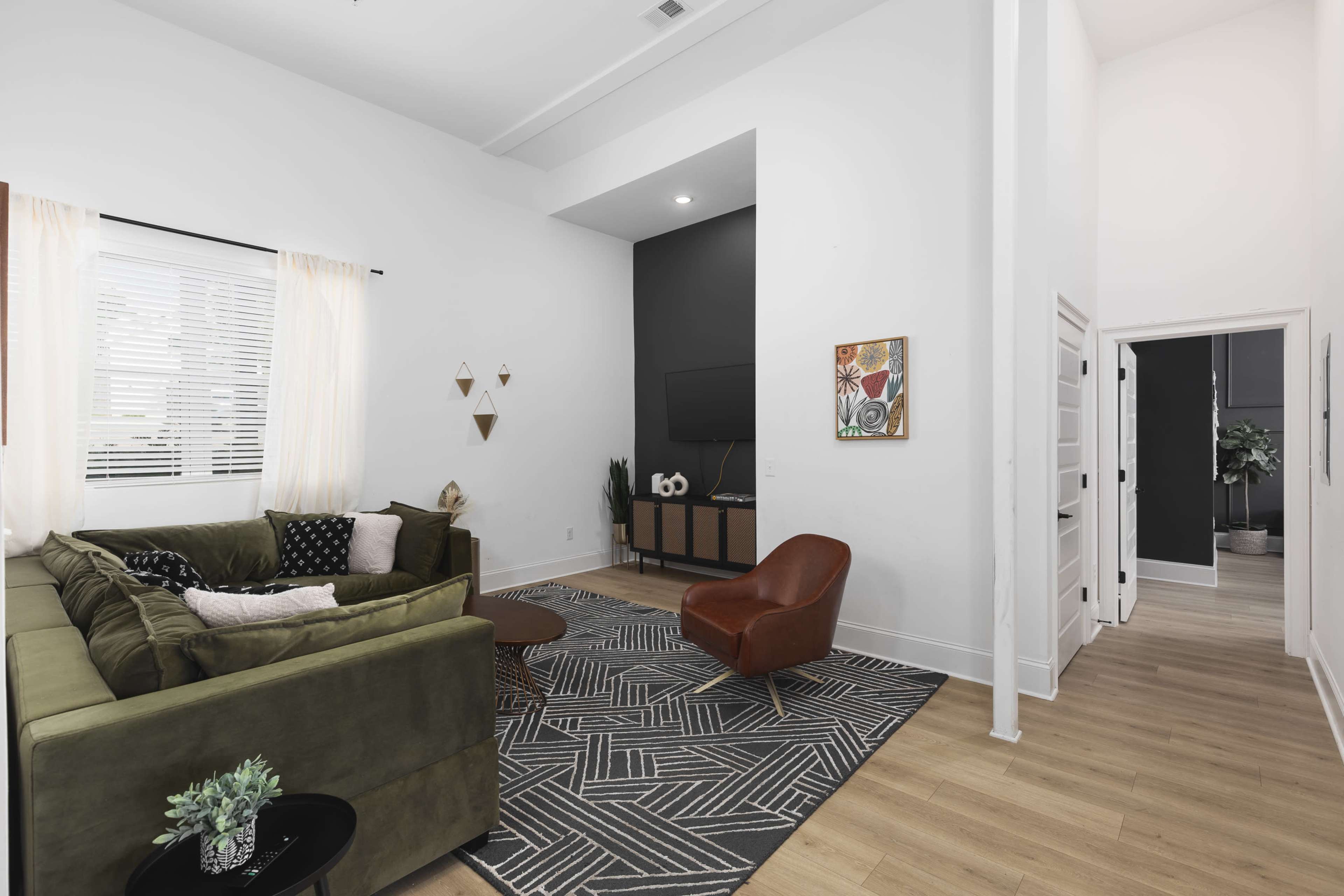 The living room features a green sectional sofa, a round coffee table, and a large television mounted on a dark accent wall, with a hallway leading to other rooms in the background.