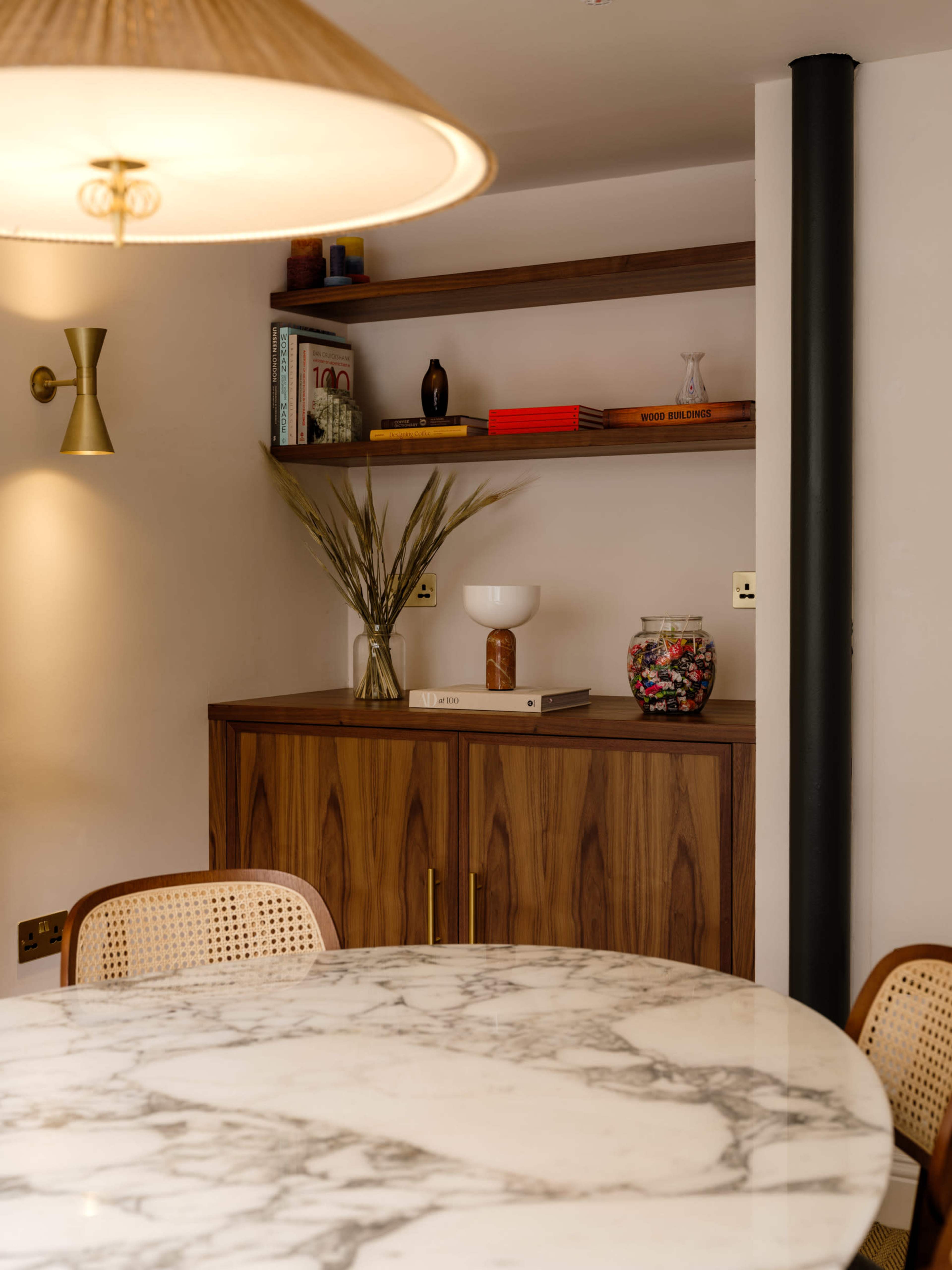 A marble dining table is set against a wooden cabinet and open shelving, displaying decorative items and plants in a well-lit room.
