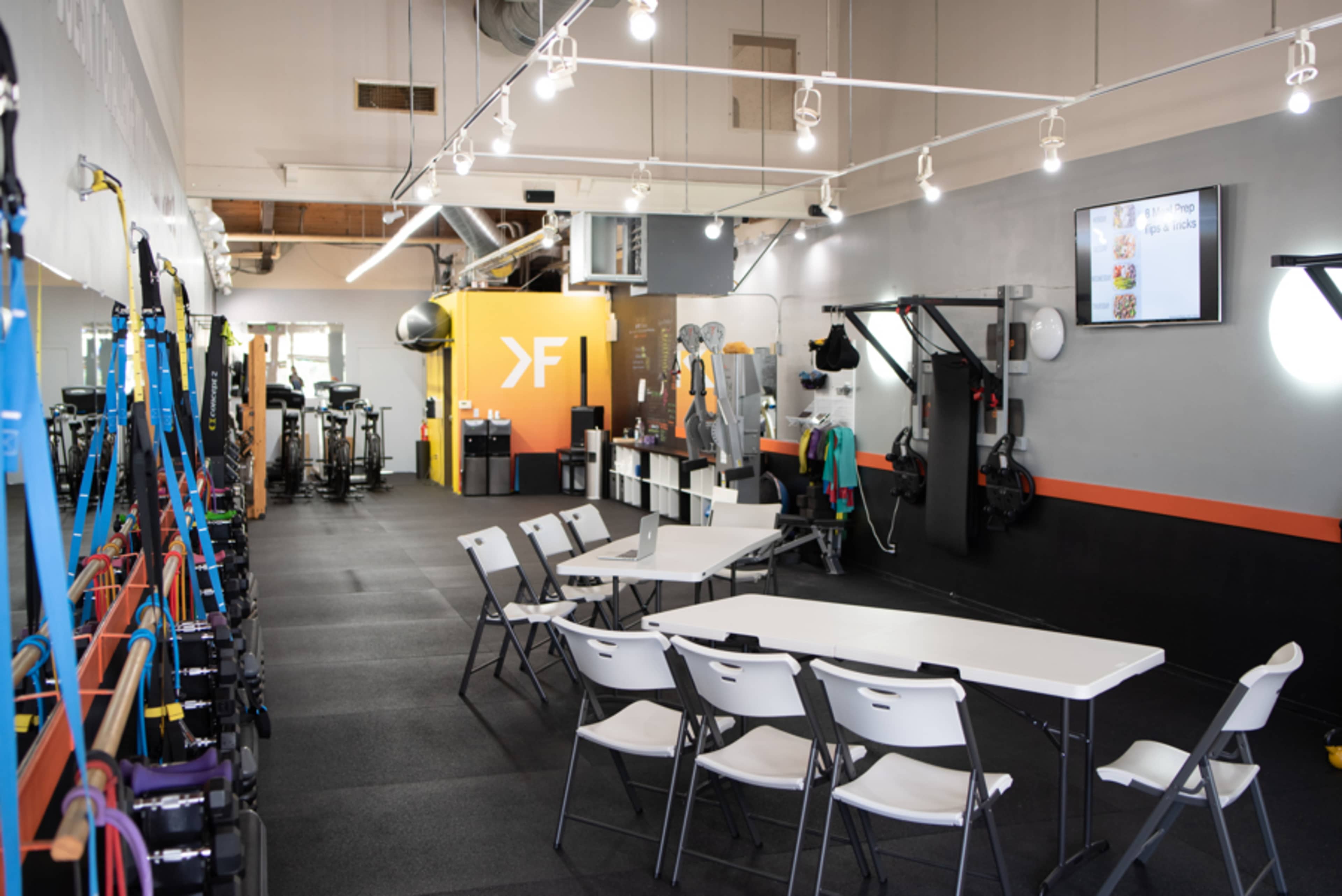 The image shows a fitness studio equipped with exercise equipment, resistance bands, and a central area featuring a table and chairs for group activities.