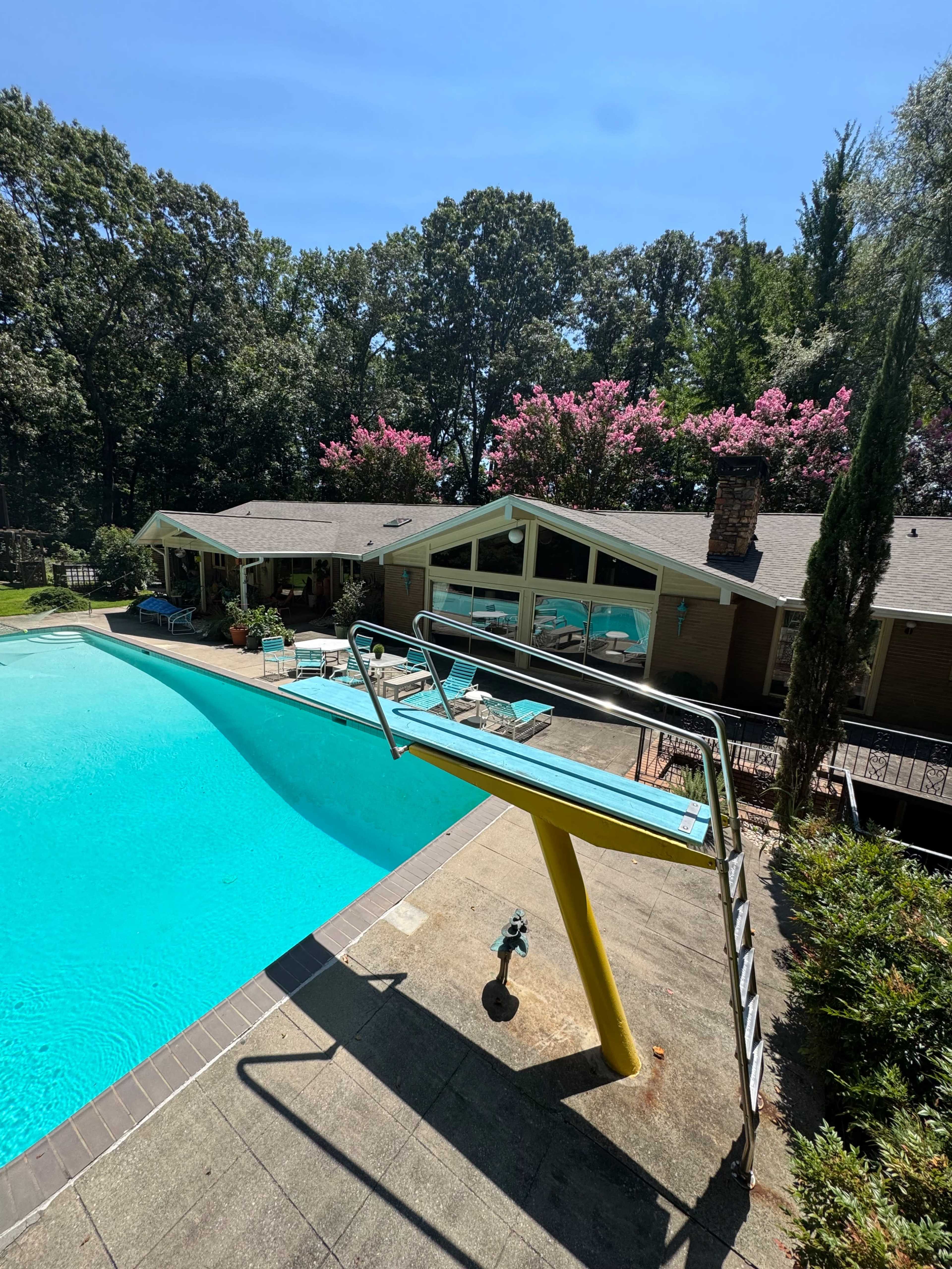 The image shows a swimming pool with a diving board on the side, surrounded by a house and landscaped trees in a sunny outdoor setting.