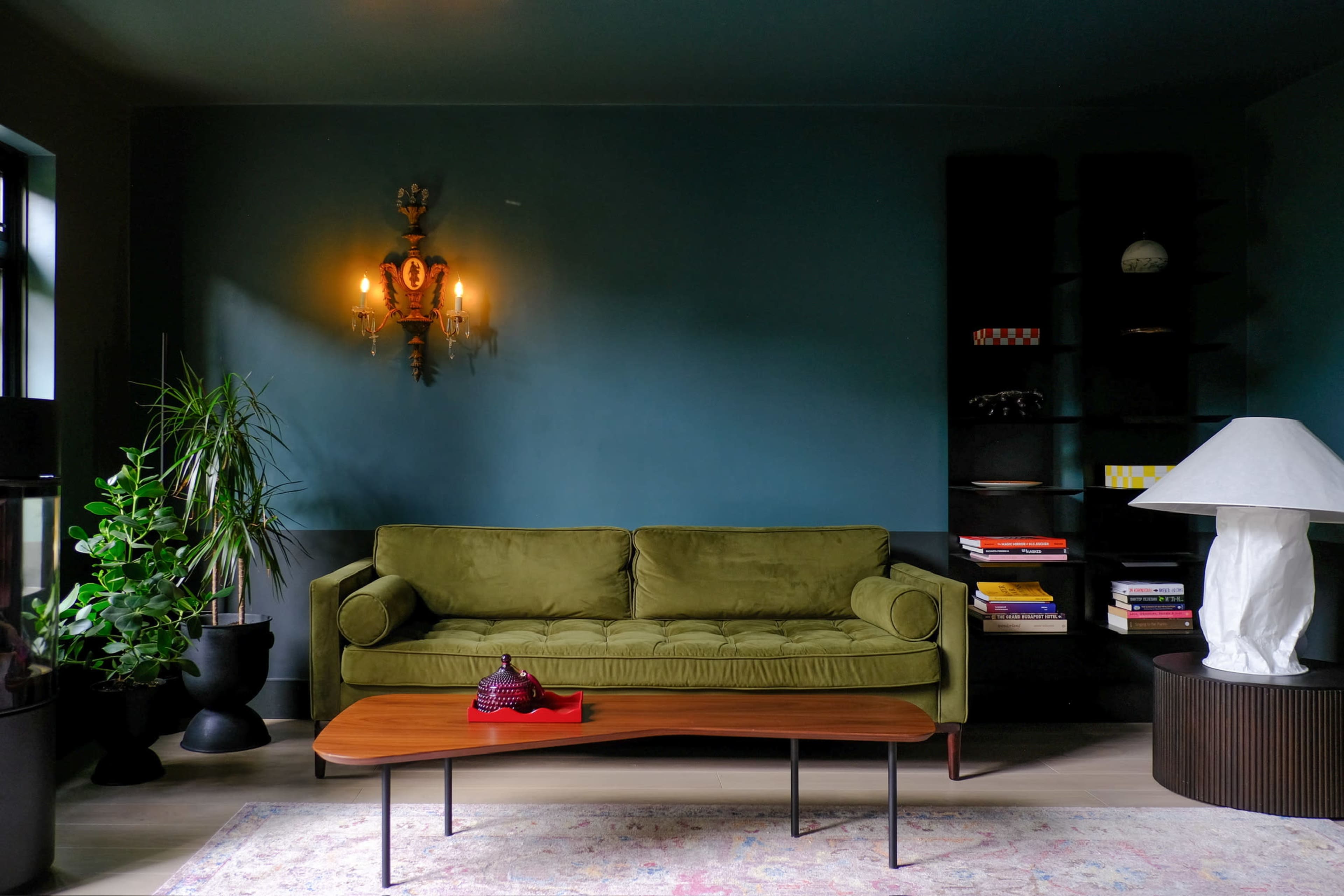A green velvet sofa is positioned against a teal wall, accompanied by a wooden coffee table, a plant in a black pot, and decorative shelves filled with books and objects.