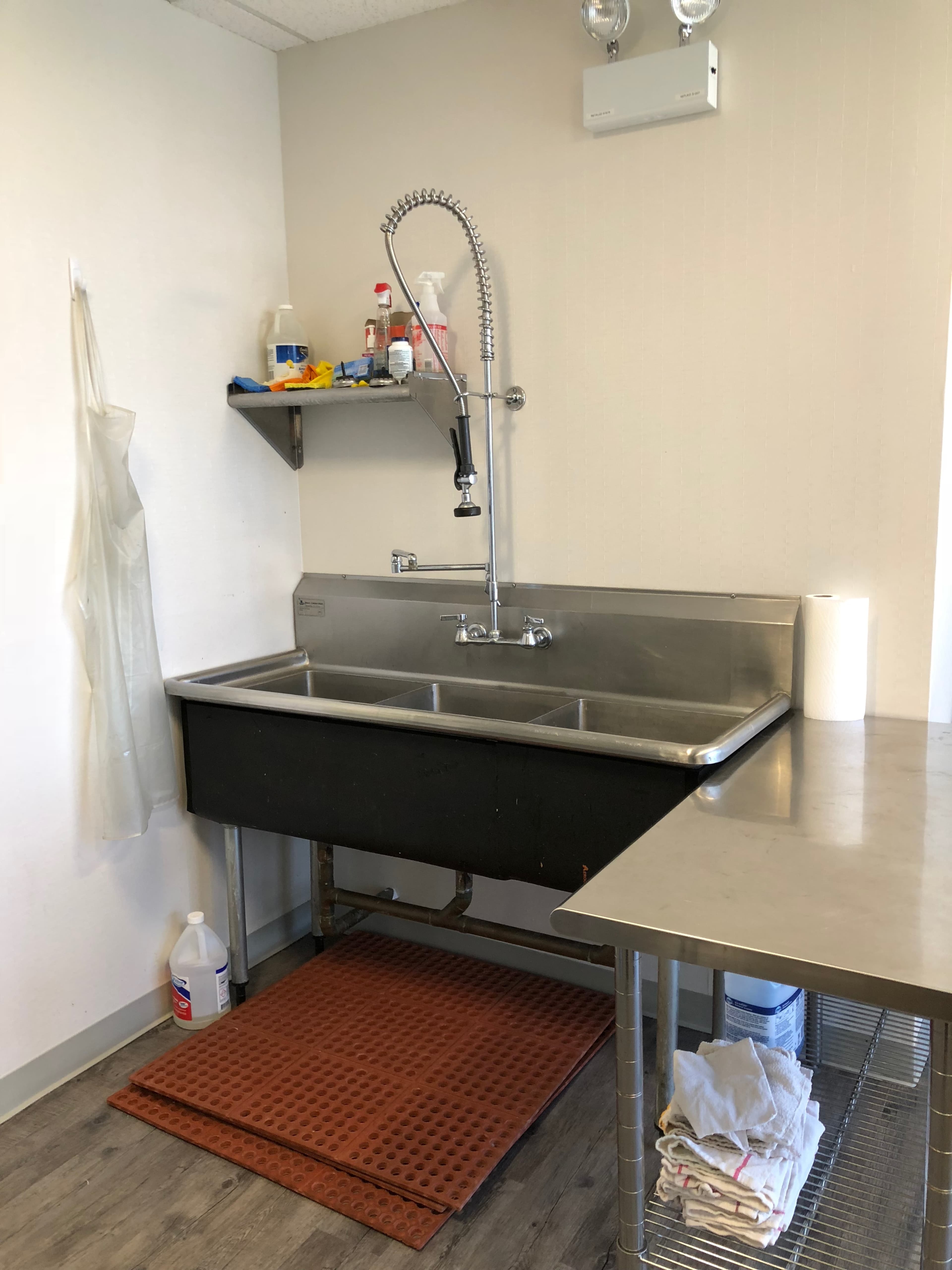 The image shows a stainless steel utility sink with a high-arch faucet in a small kitchen area, accompanied by a shelf containing cleaning supplies and a roll of paper towels.