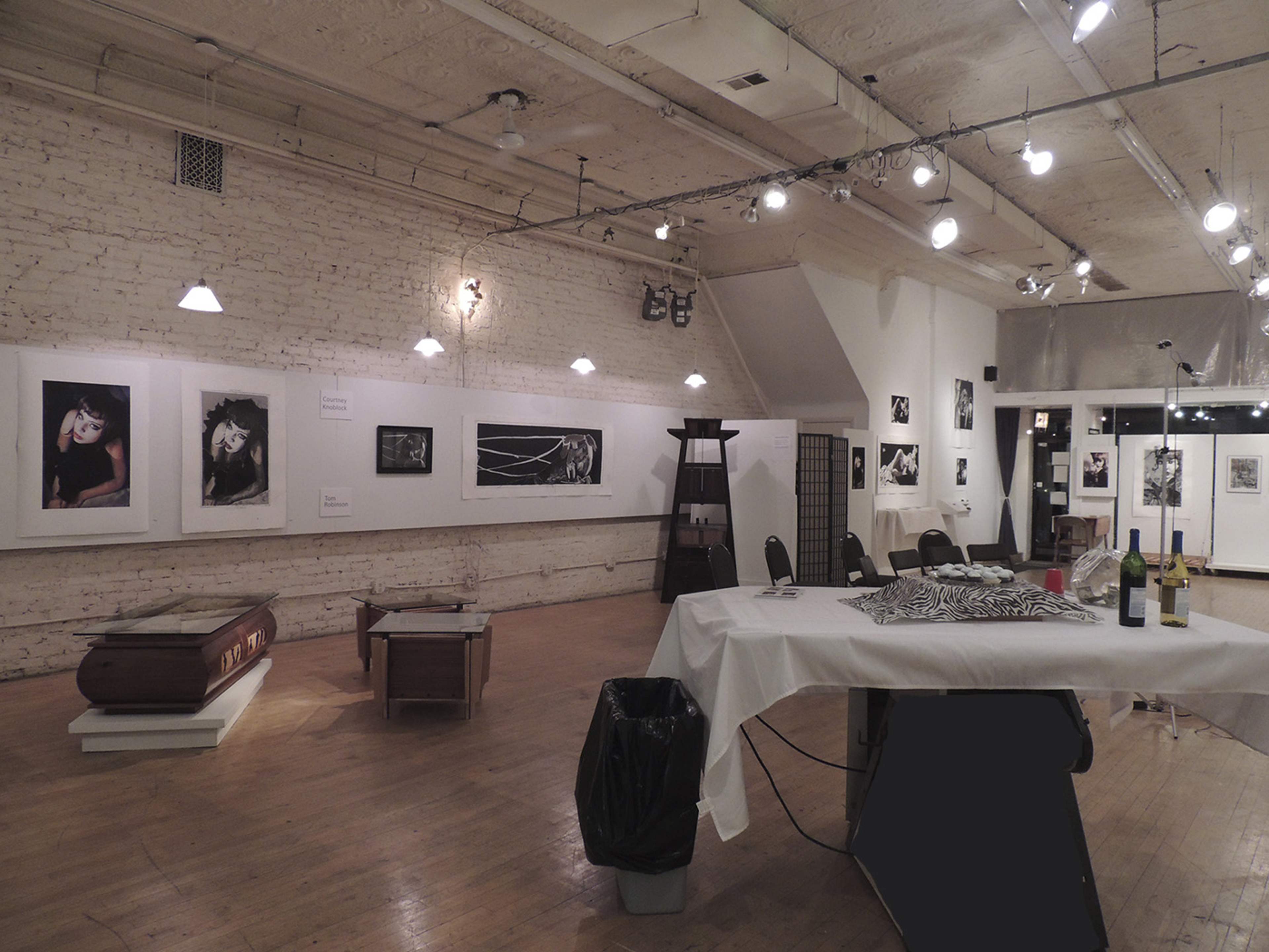 An art gallery interior featuring black and white photographs displayed on white brick walls, with a table set for a gathering and various furnishings throughout the space.