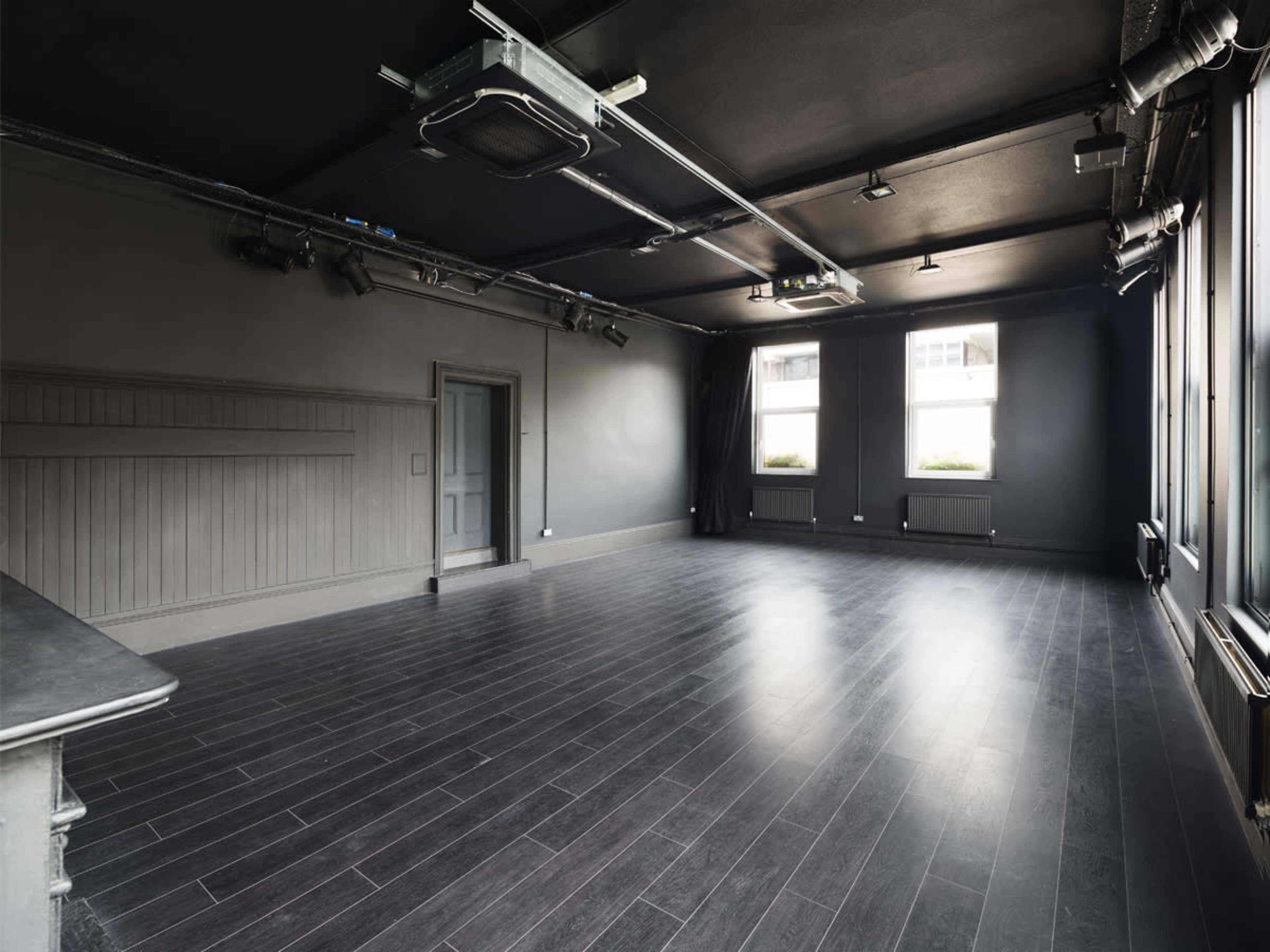 The image shows an empty room with dark walls, a wooden floor, and large windows that allow natural light to enter.