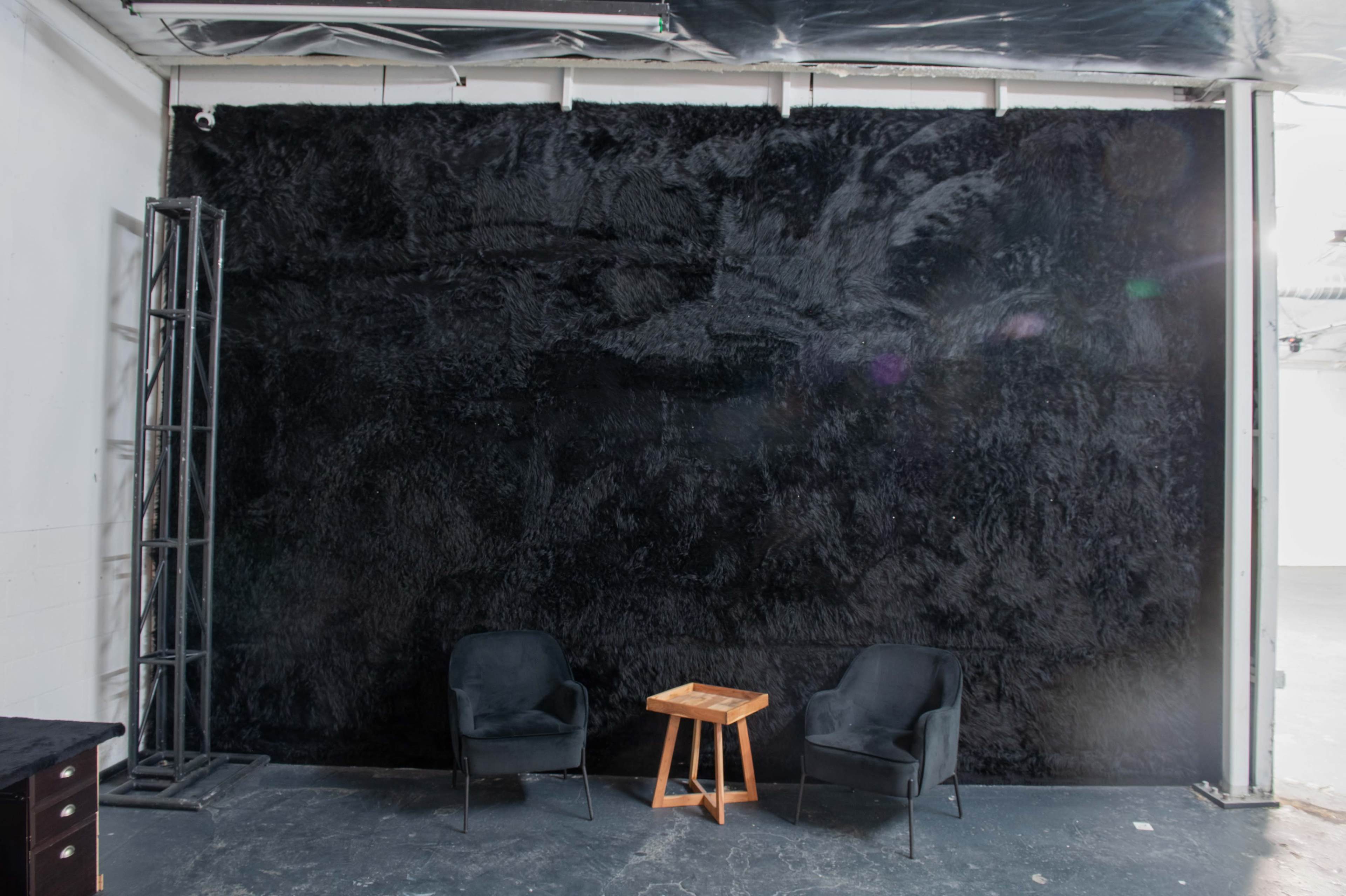 A black textured wall is positioned behind two black chairs and a small wooden table in a minimalist interior space.