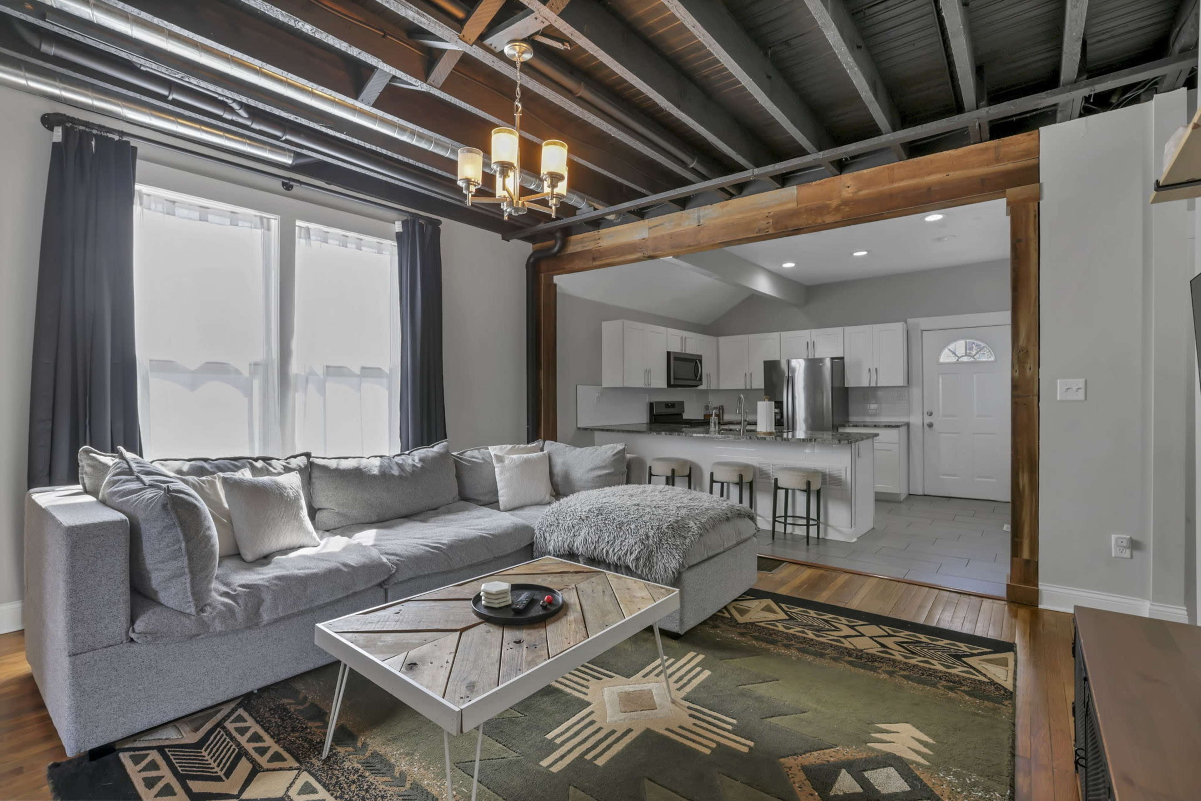 The image shows a modern living room with a gray sectional sofa, a wooden coffee table, and a kitchen area in the background featuring stainless steel appliances and white cabinetry.