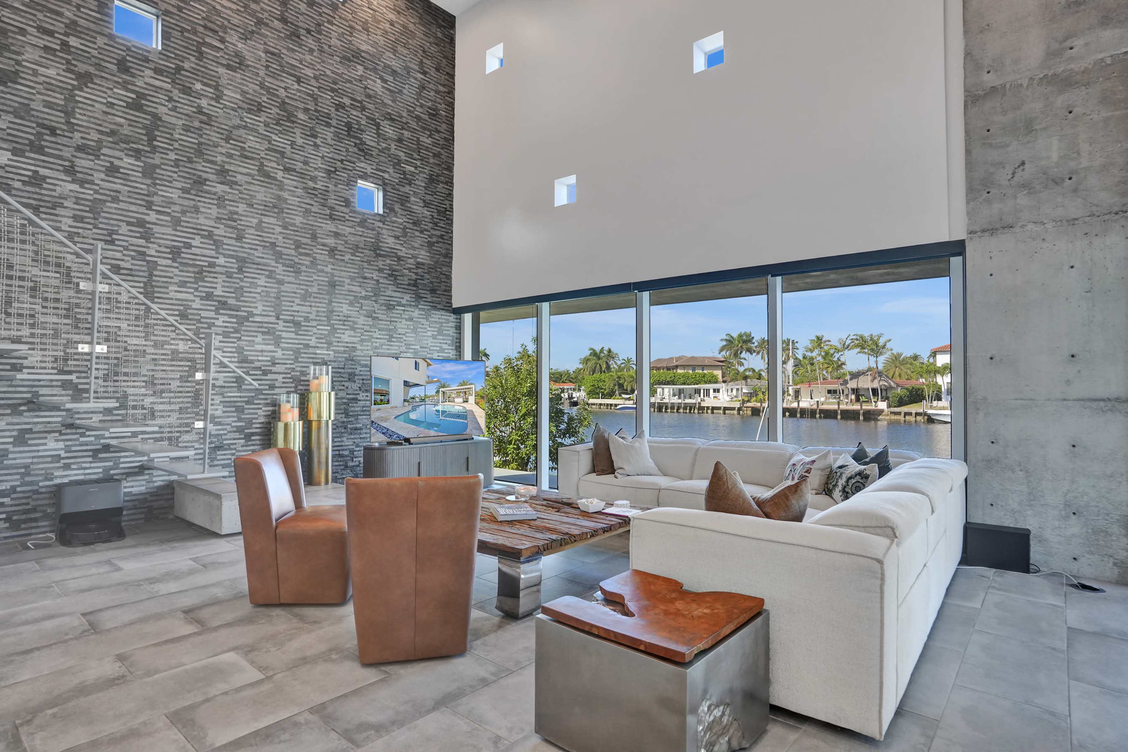 The image shows a modern living room with large windows overlooking a canal and outdoor area, featuring a sectional sofa, a wooden coffee table, and several decorative elements.