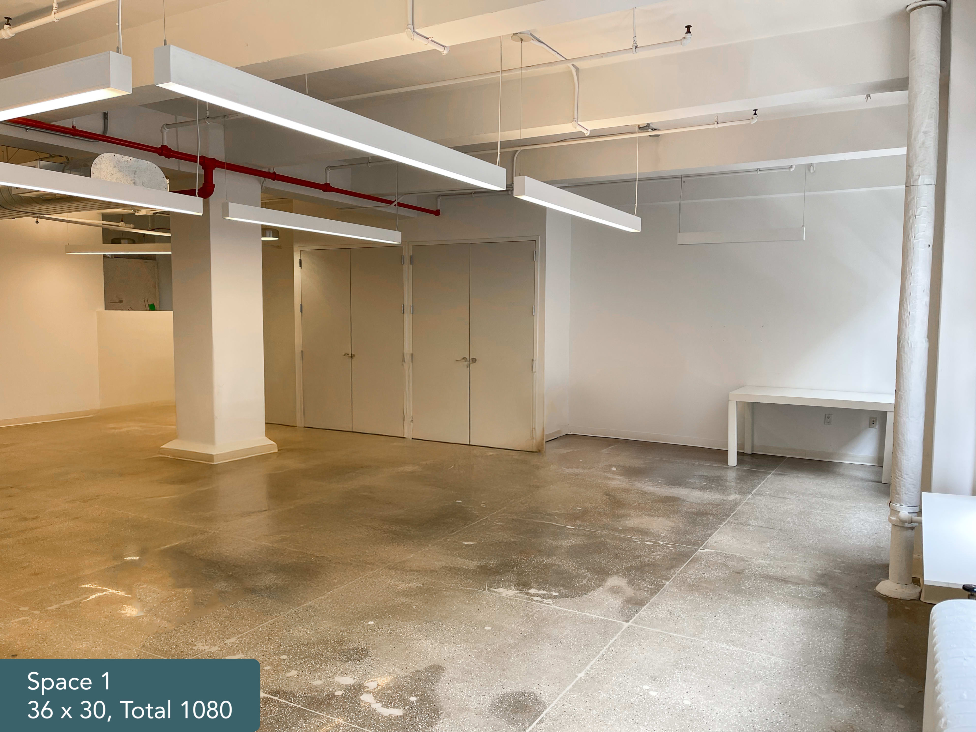 The image shows a spacious, empty room with a polished concrete floor, large ceiling lights, and a white wall with two double doors.