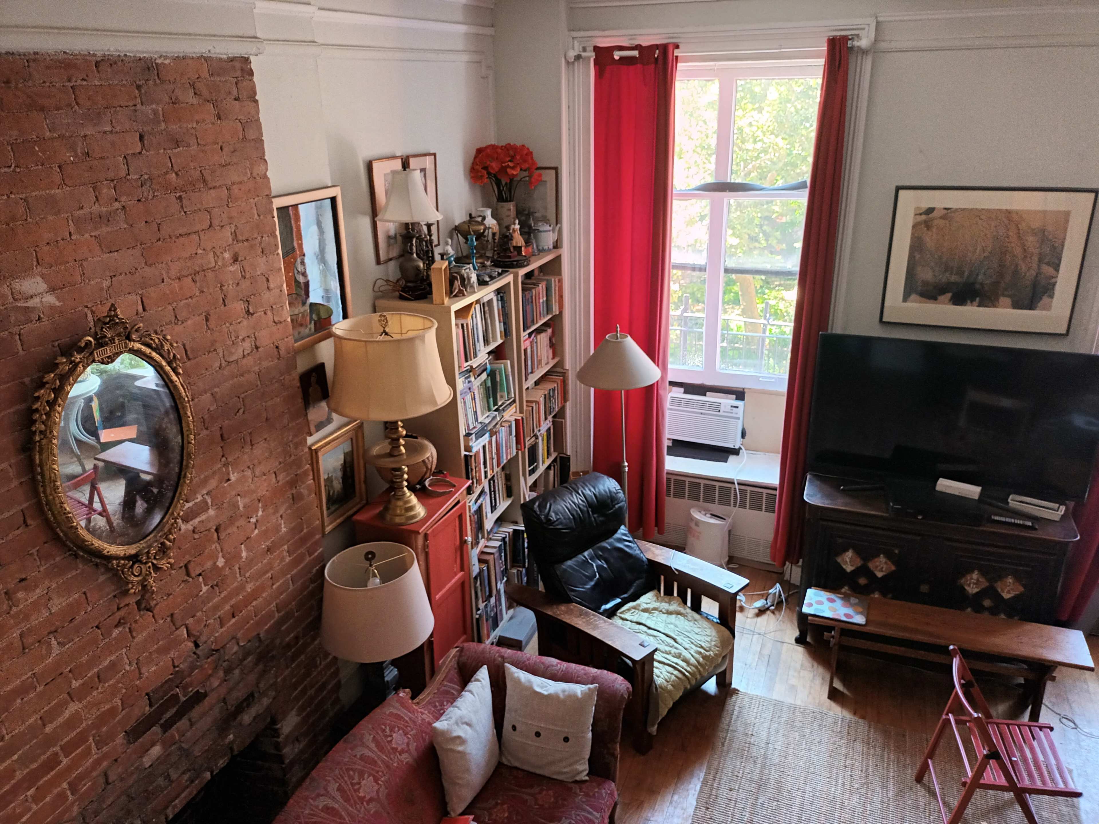 A cozy living room with a brick wall, a bookshelf filled with books, a television, a red sofa, and large windows allowing natural light to enter.