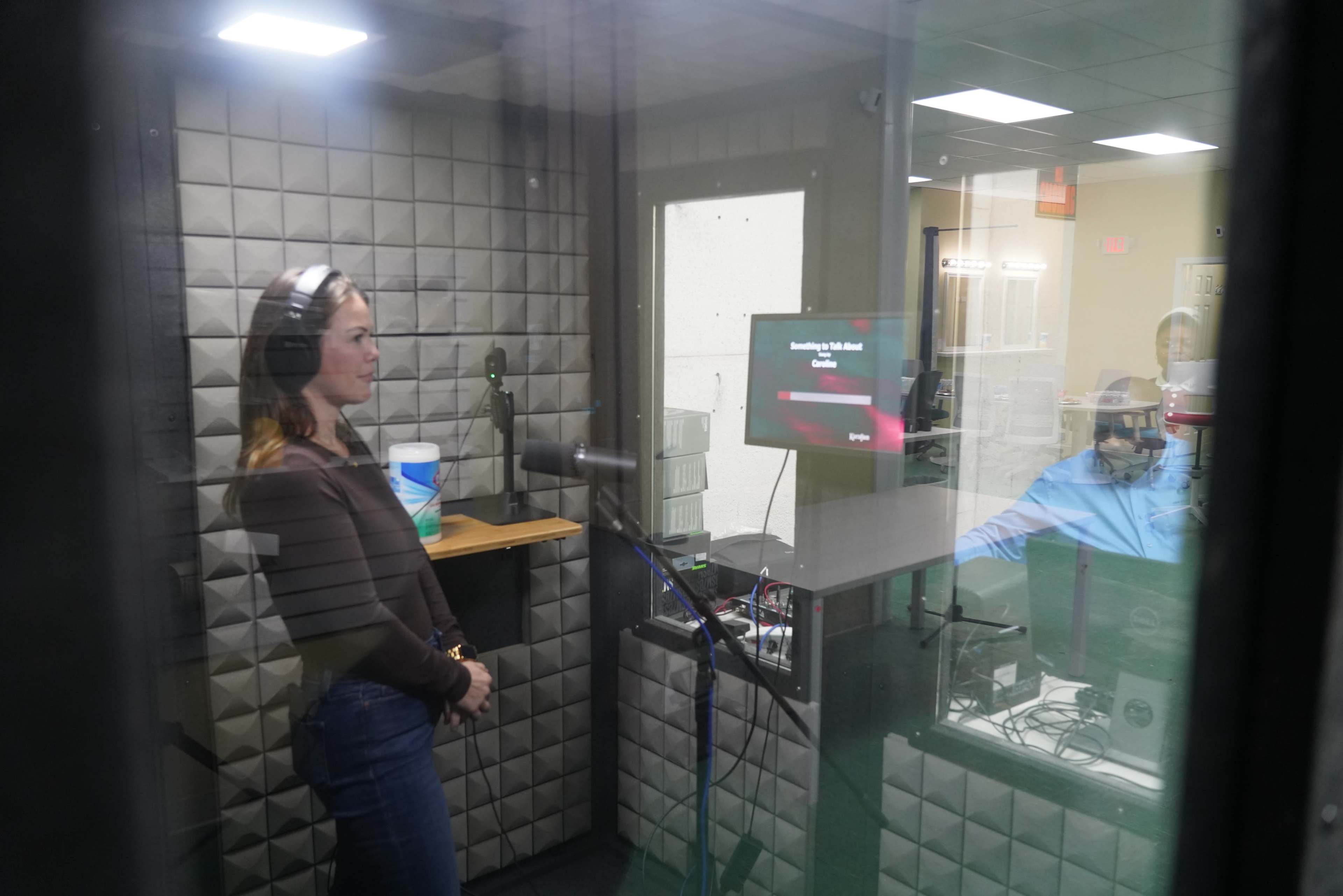 A woman is standing in a soundproof booth wearing headphones while a person sits at a desk with a monitor in front of them.