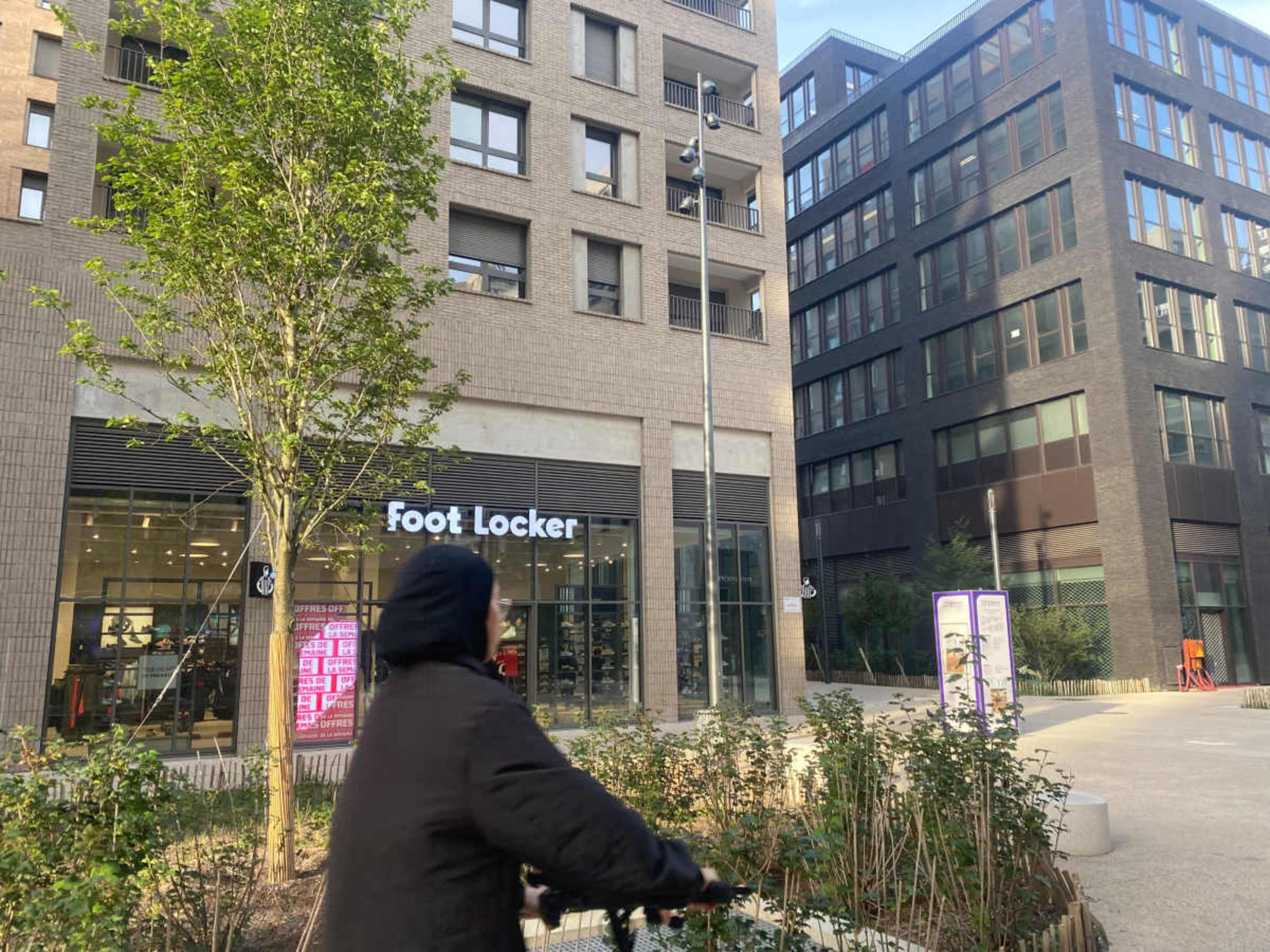 A person on a bicycle passes by a Foot Locker store located in a modern urban area with brick buildings.