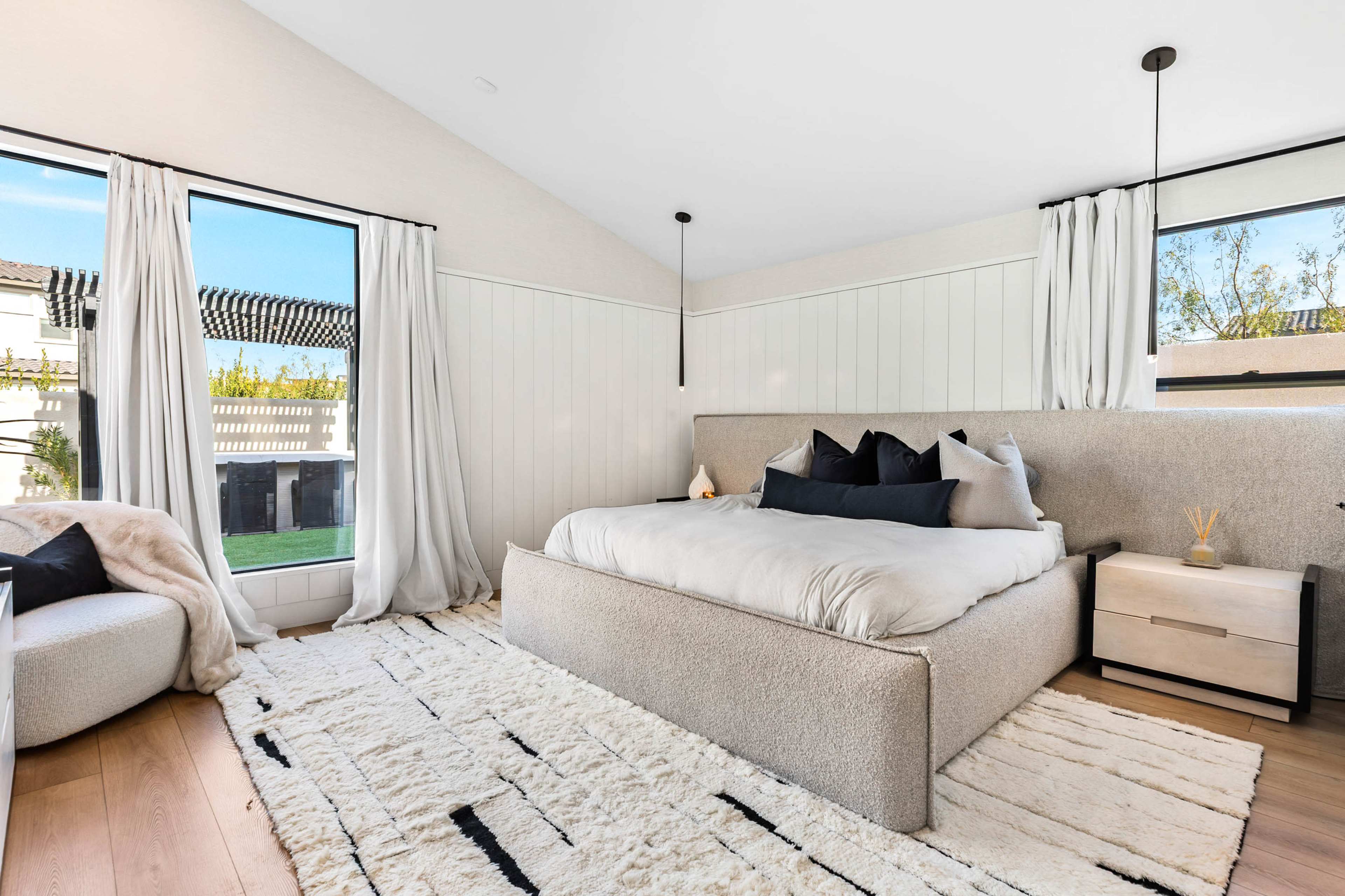 The image depicts a modern bedroom featuring a large bed with a light-colored comforter, a rug with a geometric pattern, and large windows allowing natural light to enter.