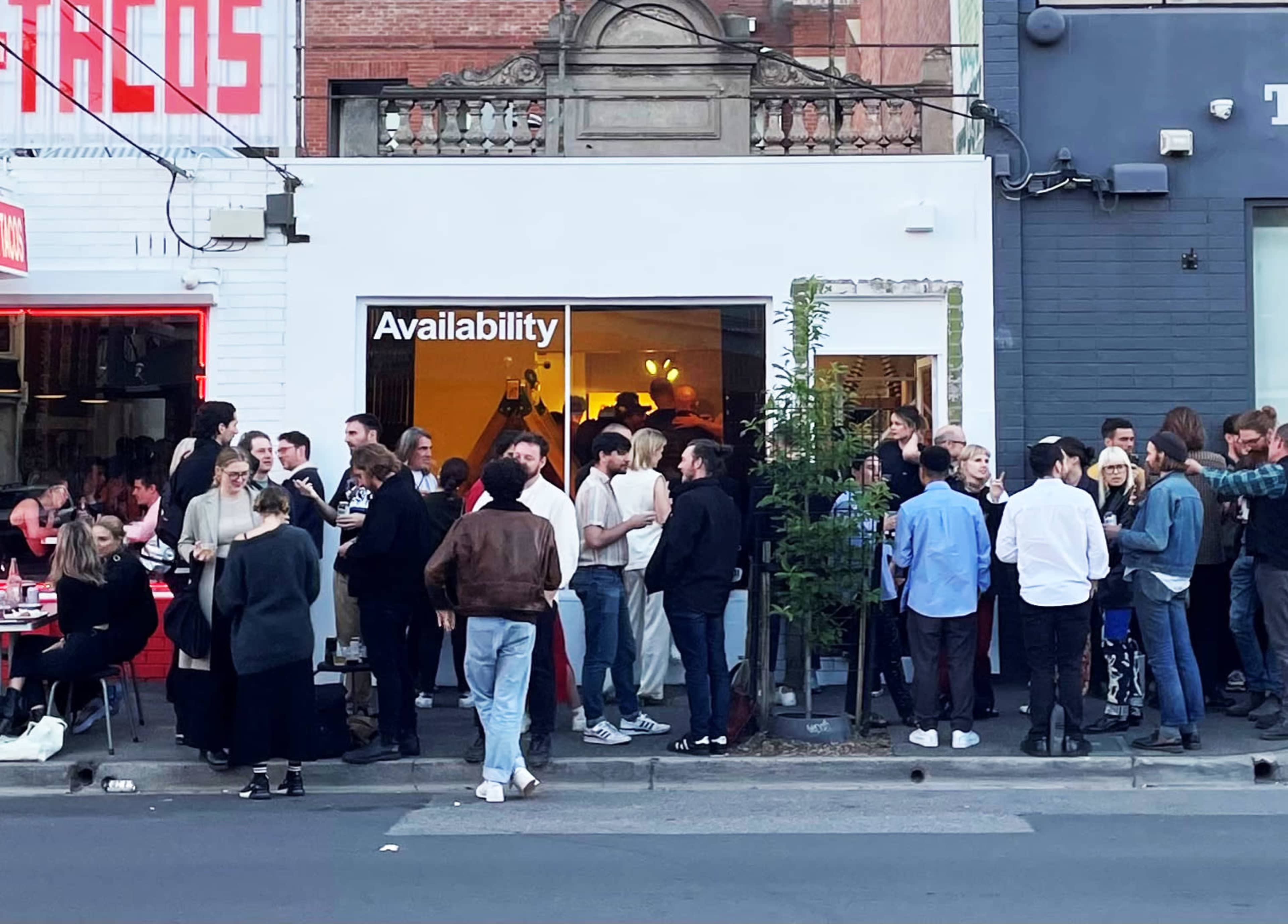 A crowd of people gathers outside a shop labeled "Availability," with people standing and socializing on the sidewalk.
