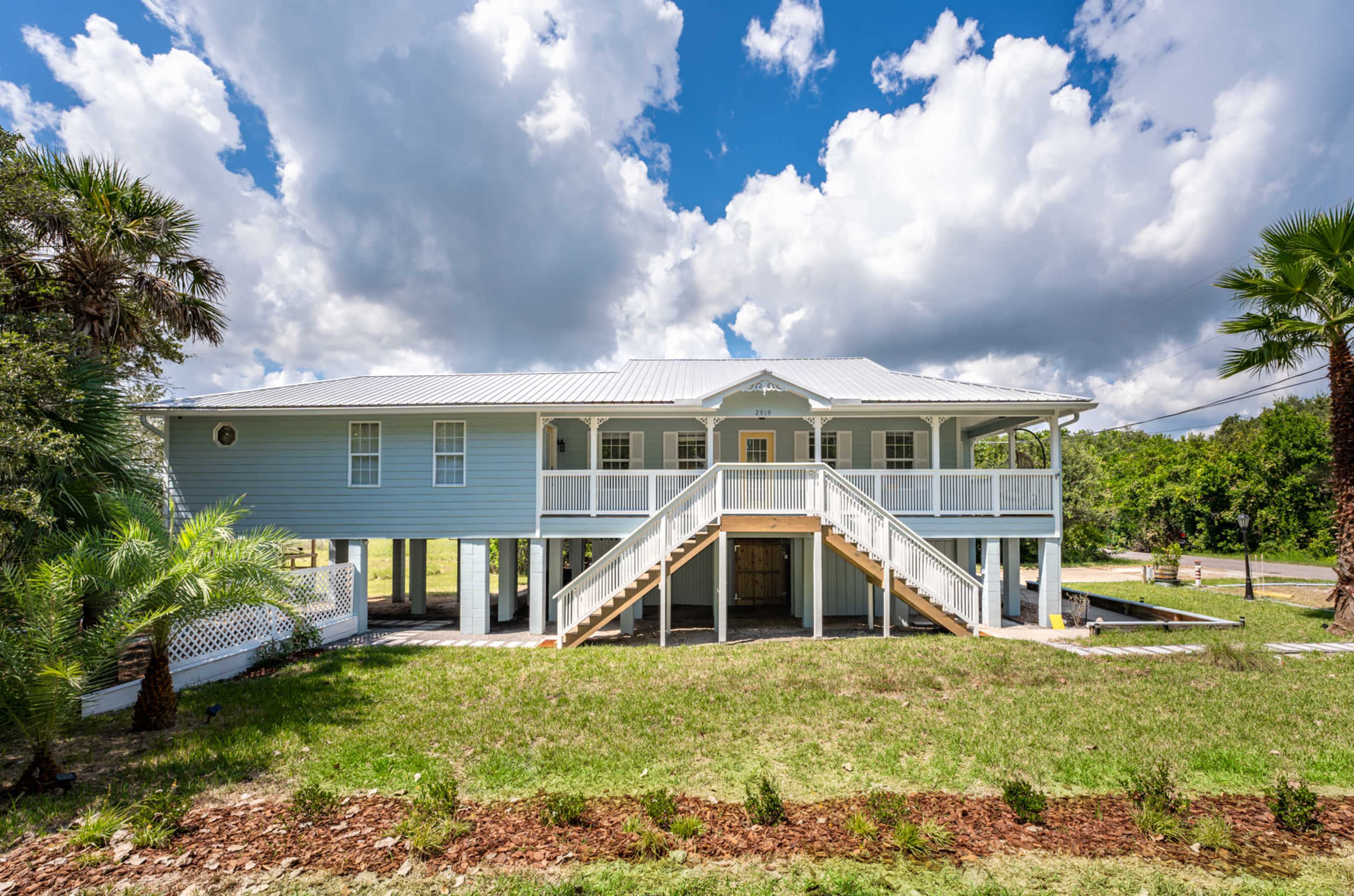 A two-story blue house with a metal roof features a wide front staircase and is surrounded by palm trees and a grassy yard under a partly cloudy sky.