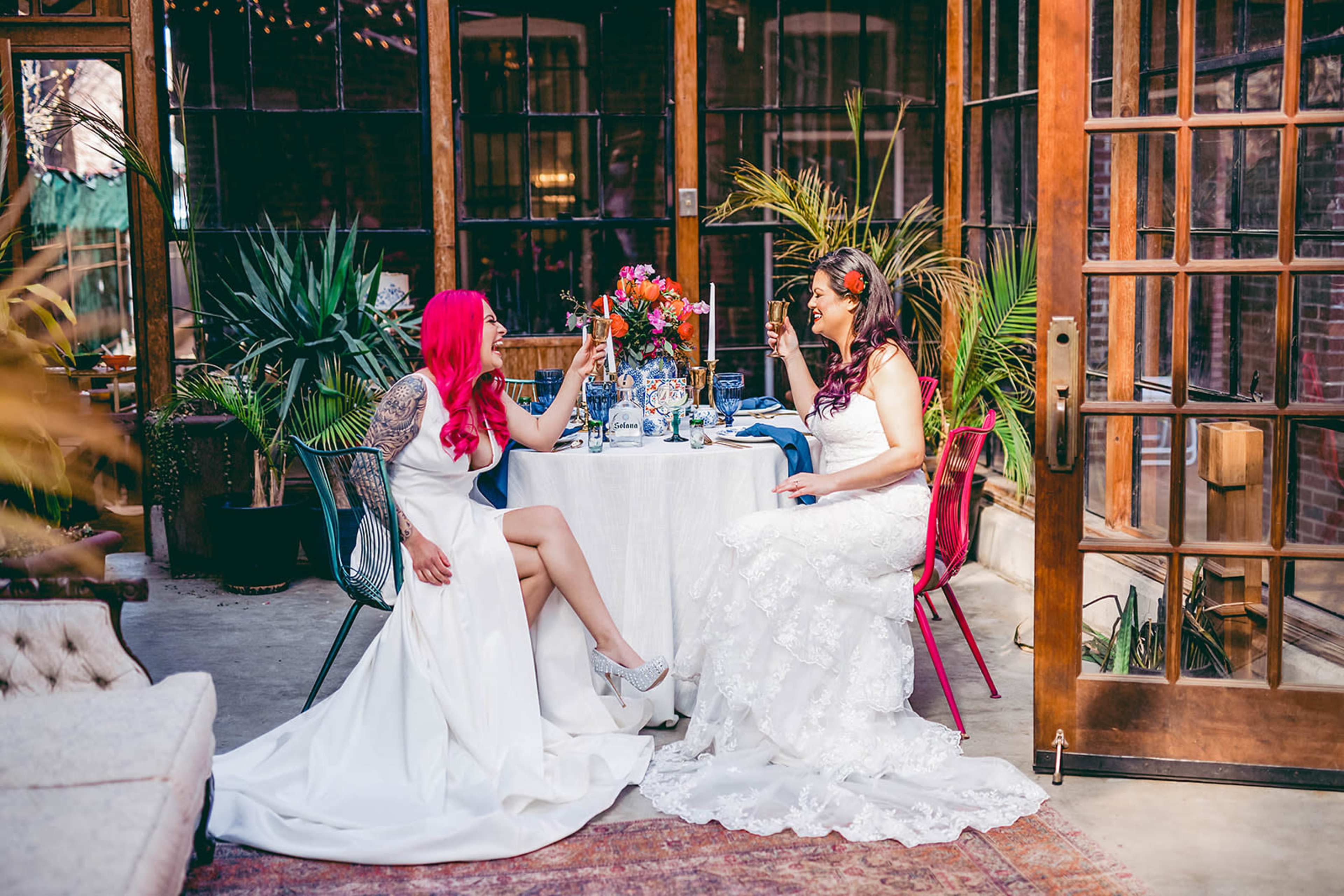 Two women sit at a decorated table with flowers and candles in a glass-enclosed room surrounded by plants.