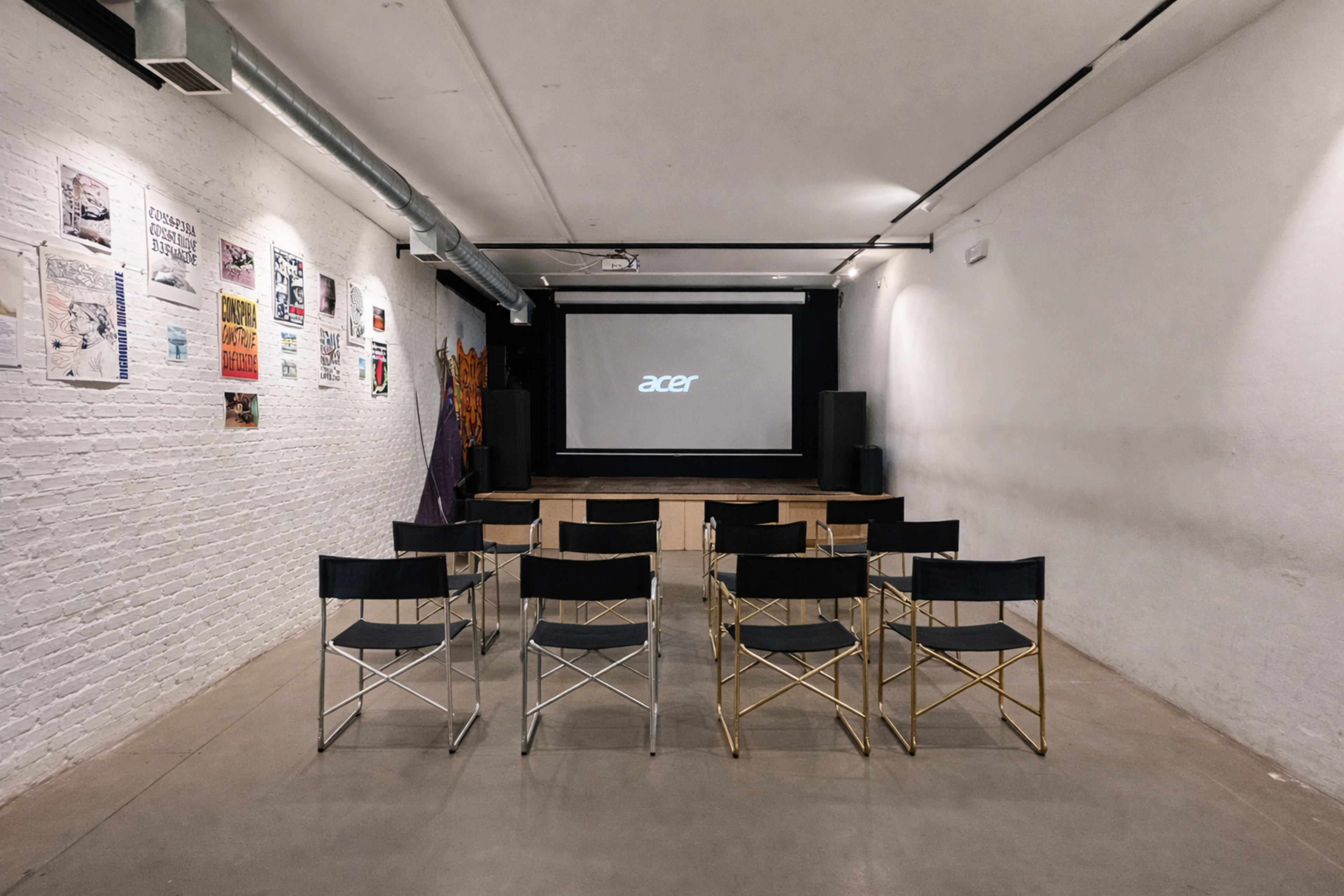 The image shows a minimalist presentation space with rows of black chairs facing a screen displaying the brand "acer," alongside posters on the left wall.