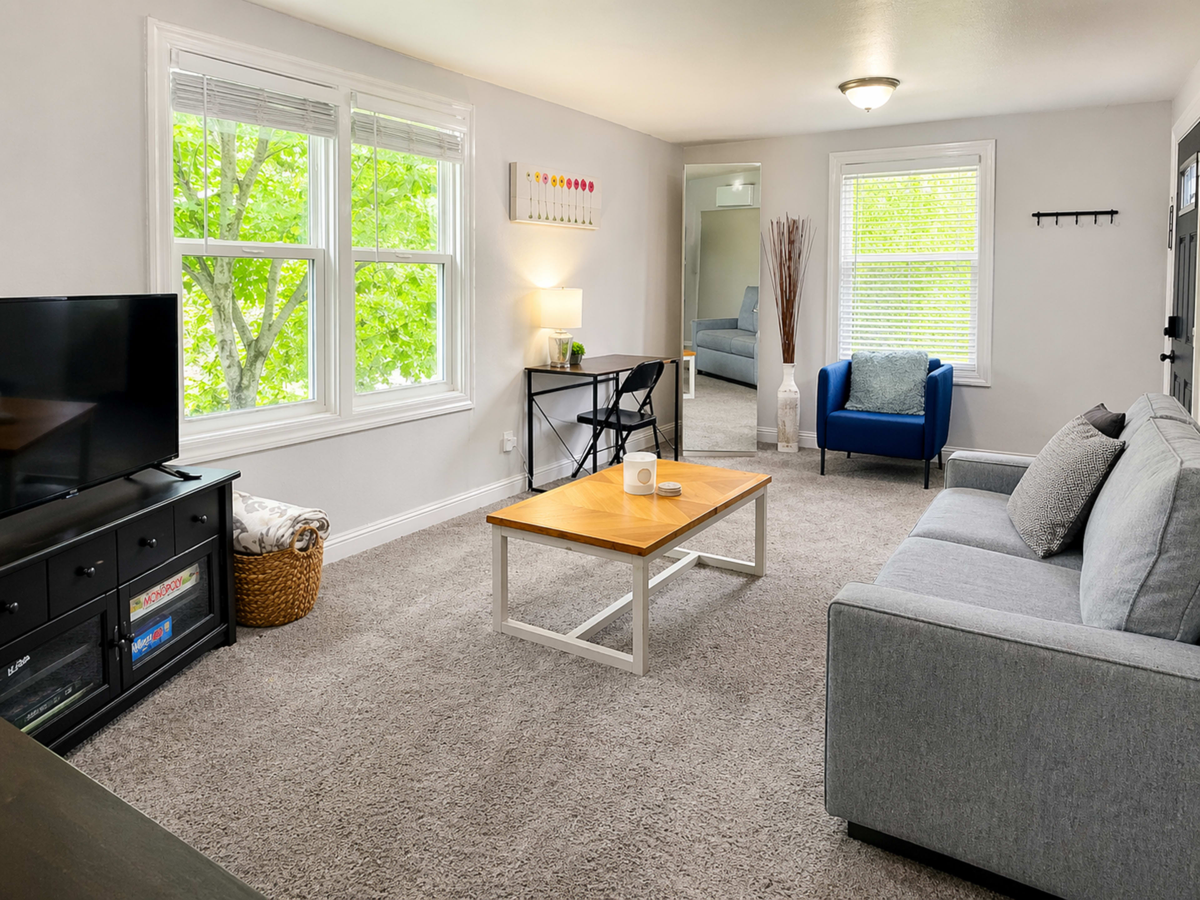 The image shows a well-lit living room featuring a gray sofa, a wooden coffee table, a television on a black cabinet, and a blue chair near a large window.