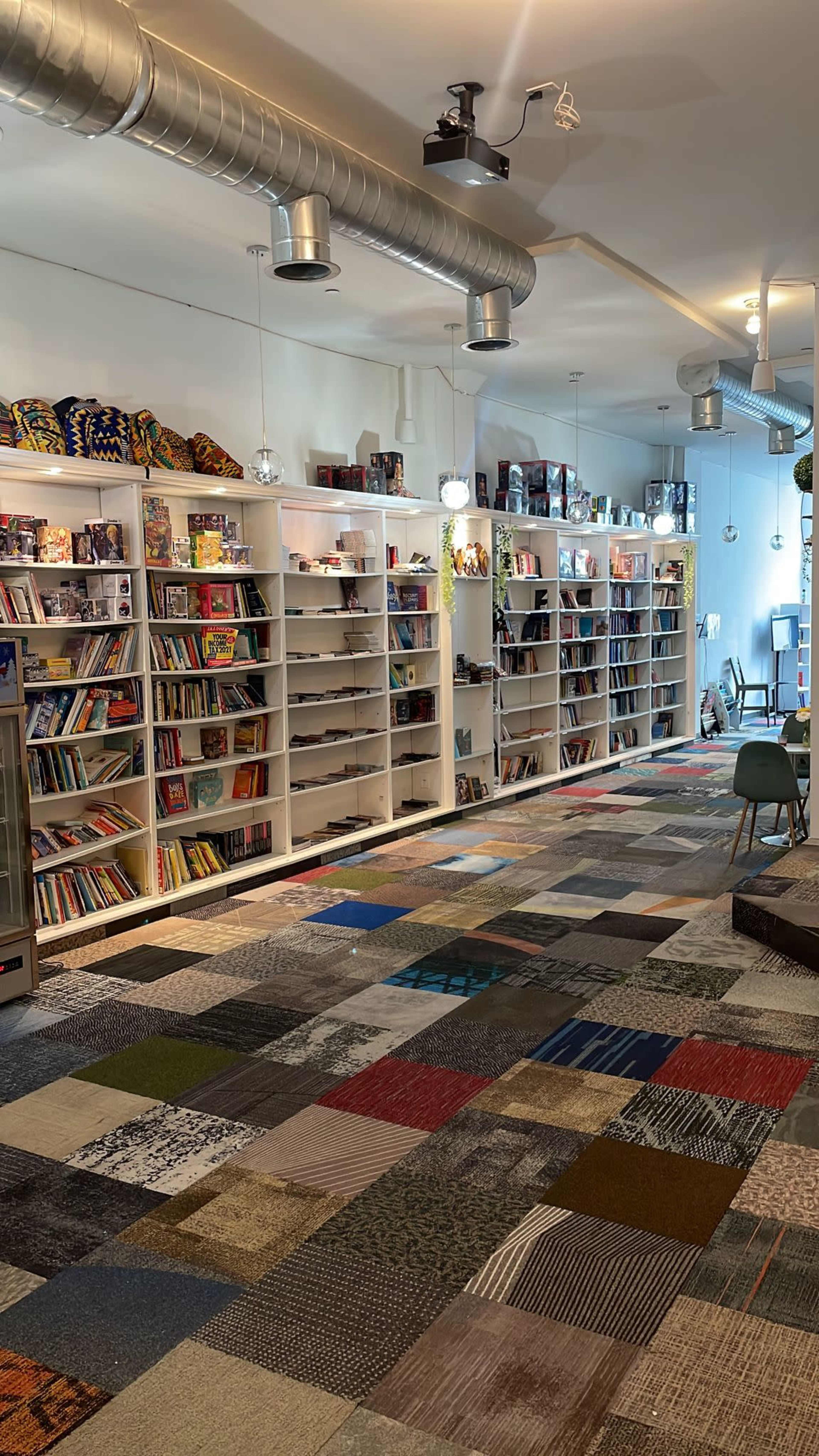 A spacious interior features brightly lit shelves filled with books and games, complemented by a colorful patchwork carpet.