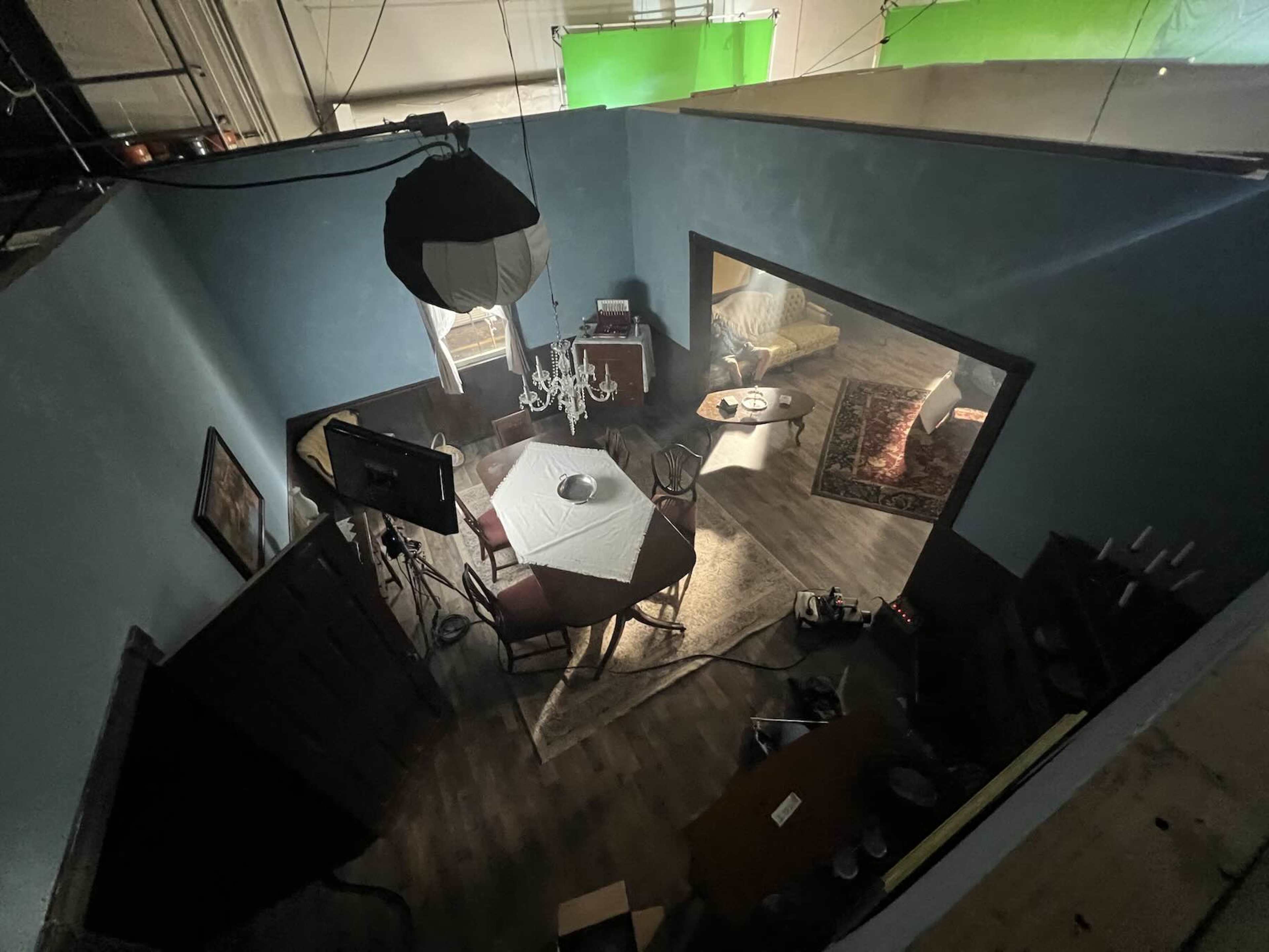 The image shows an overhead view of a dimly lit room set up for a scene, featuring a dining table with chairs, a chandelier, and a couch with a rug.