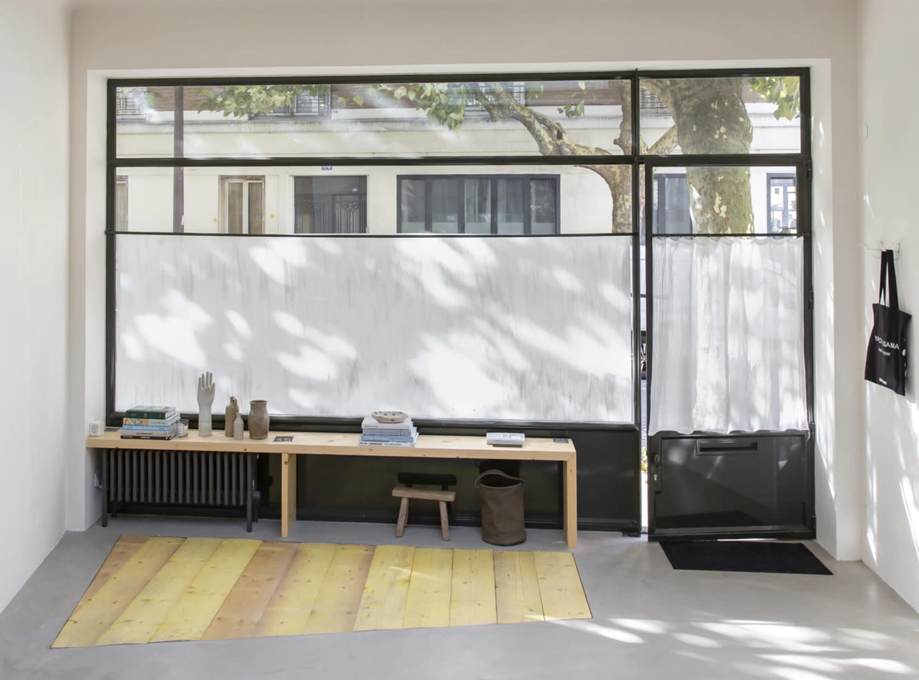 The image shows a minimalist interior space with a wooden bench displaying items, a section of wood flooring, and a large window with sheer curtains allowing natural light to enter.